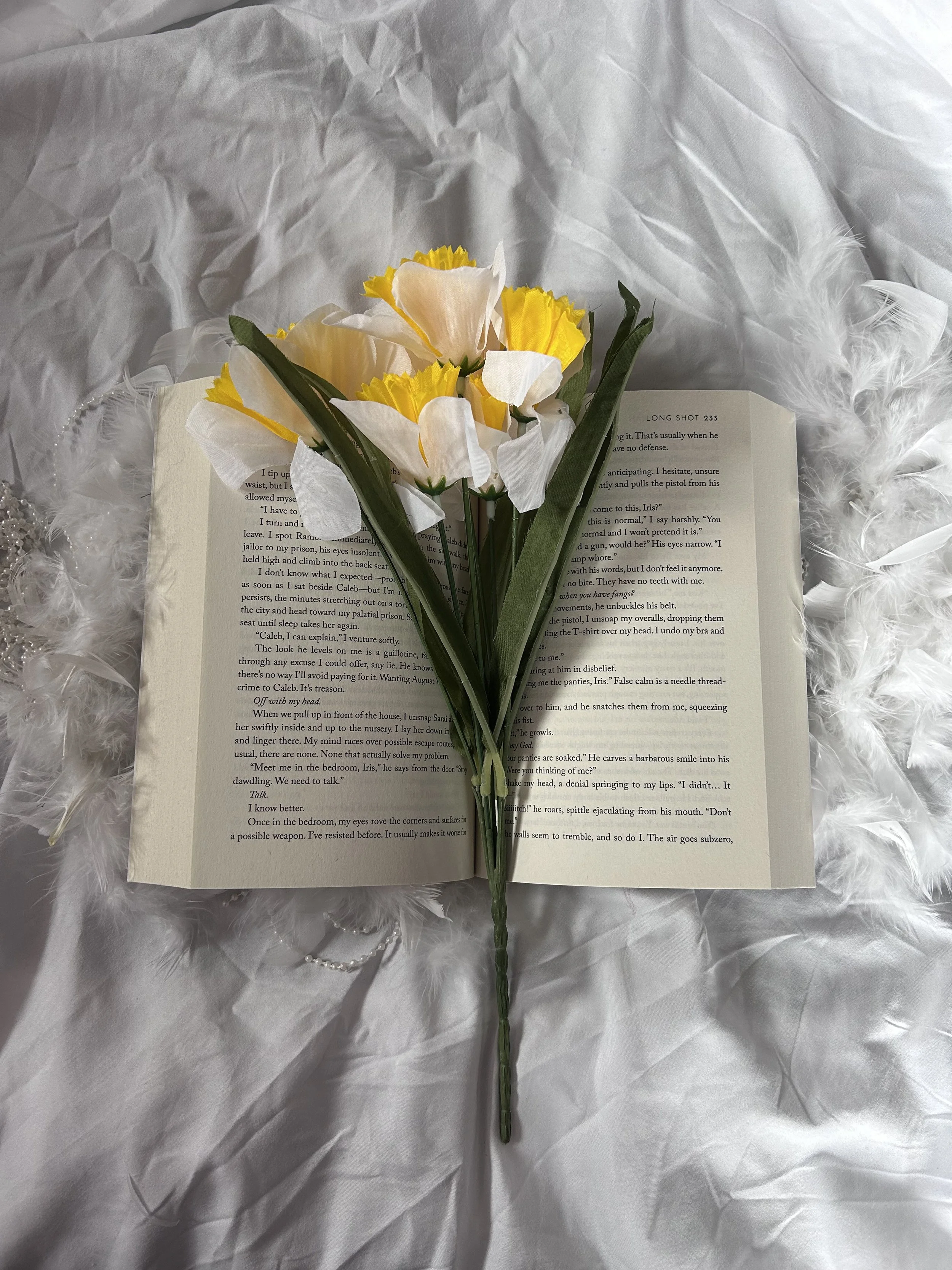 An open book with a bouquet of white and yellow flowers placed on top, resting on a white, wrinkled fabric background with a feathered accessory.
