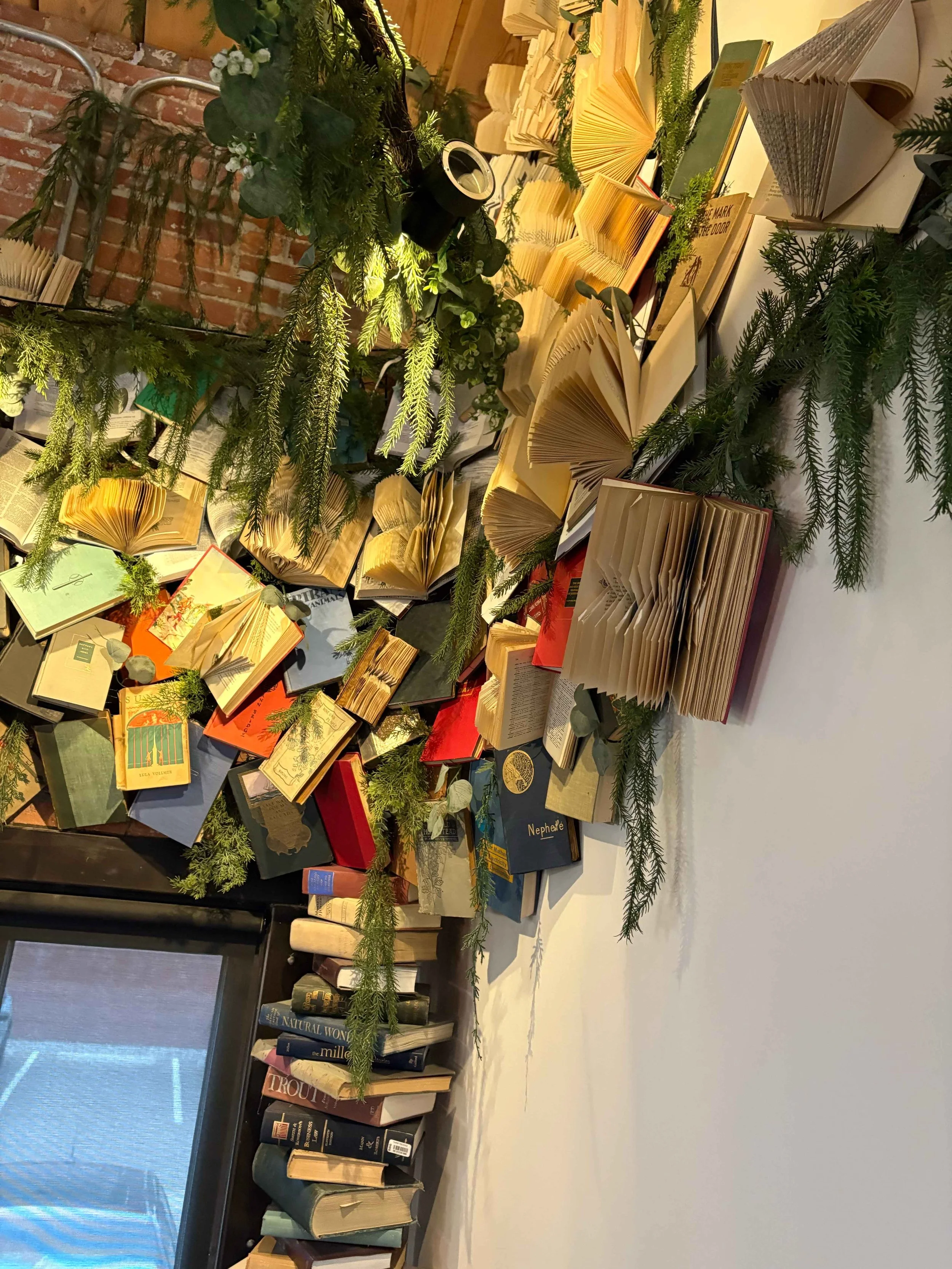 Decorative wall display of open books and greenery, including pine branches and eucalyptus leaves, in a cozy interior setting.
