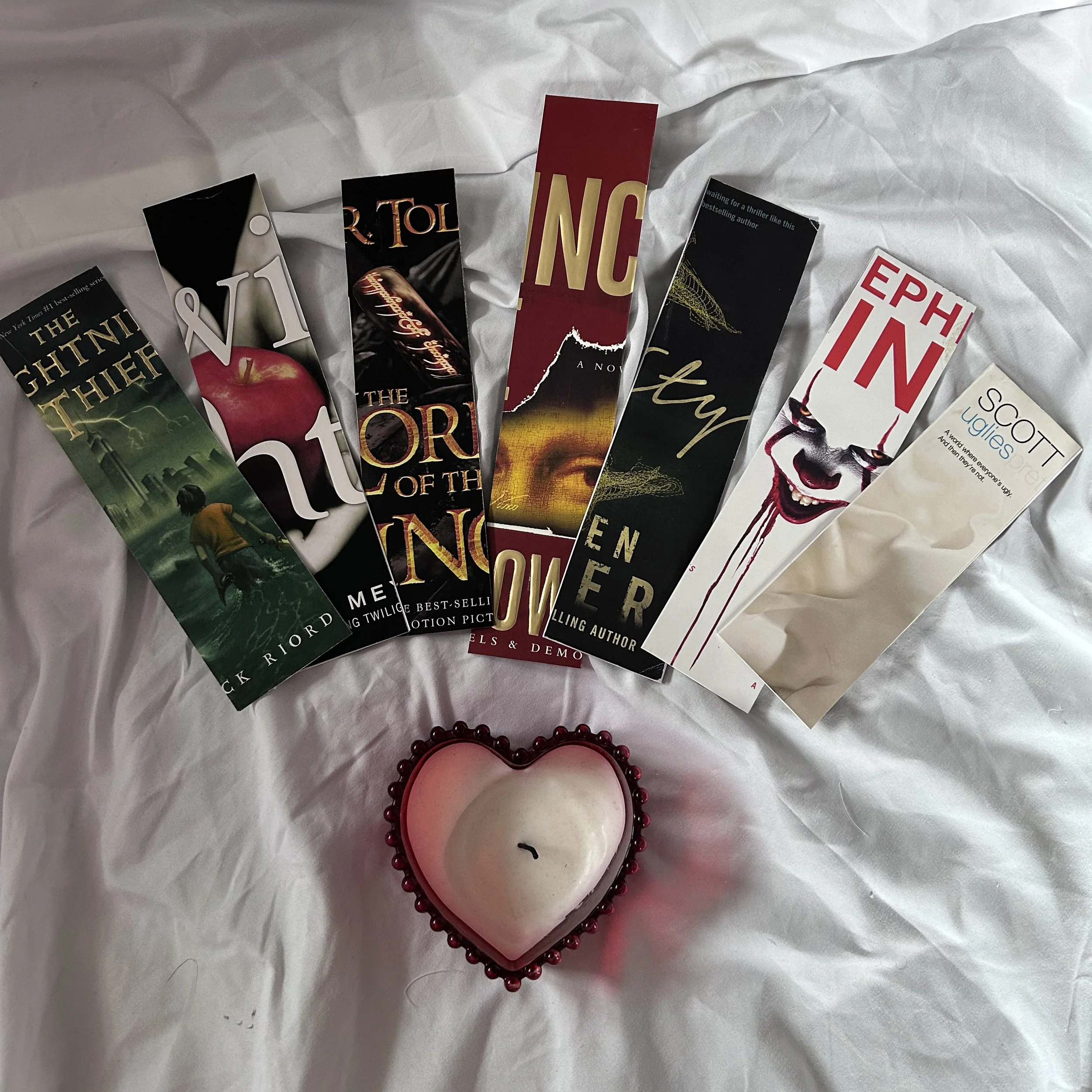 Seven bookmarks with recycle book titles / covers, and a heart-shaped candle with a beaded red border, placed on white fabric.