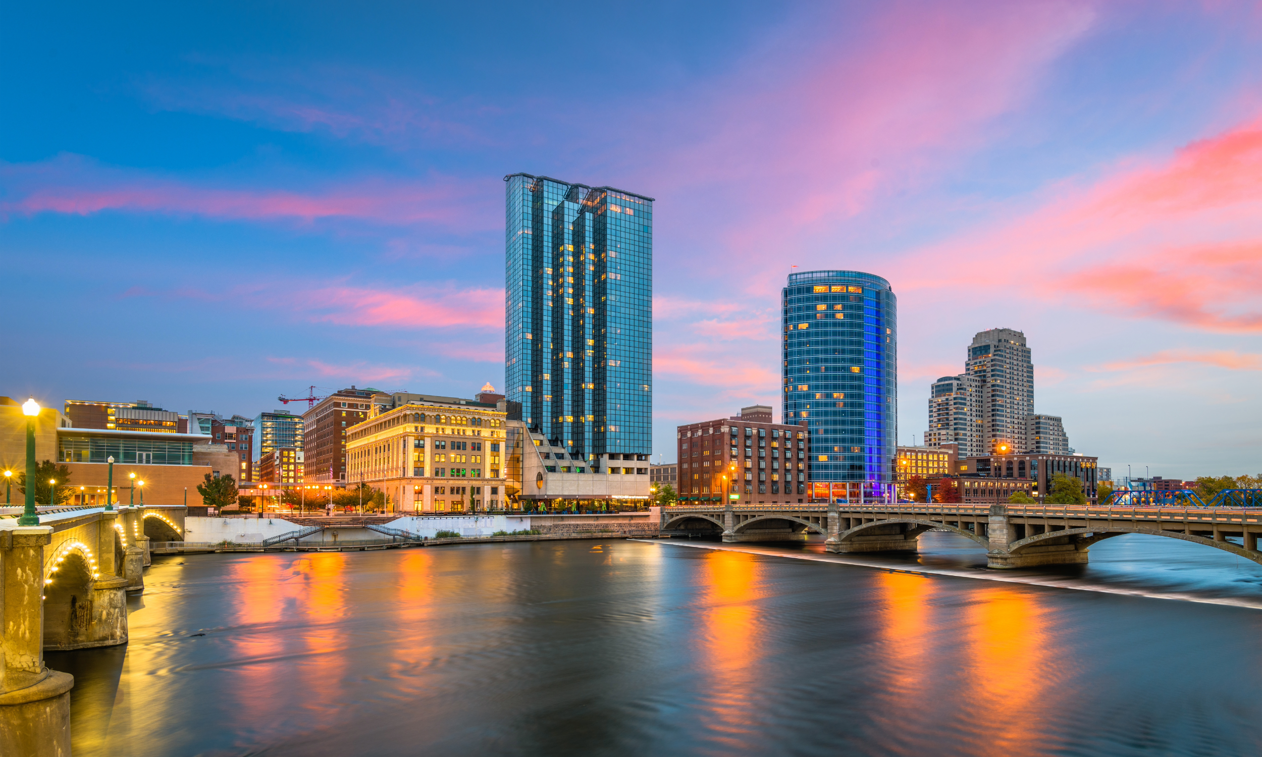 City skyline at sunset with modern glass skyscrapers, a river reflecting colorful sky, and illuminated bridge and buildings.