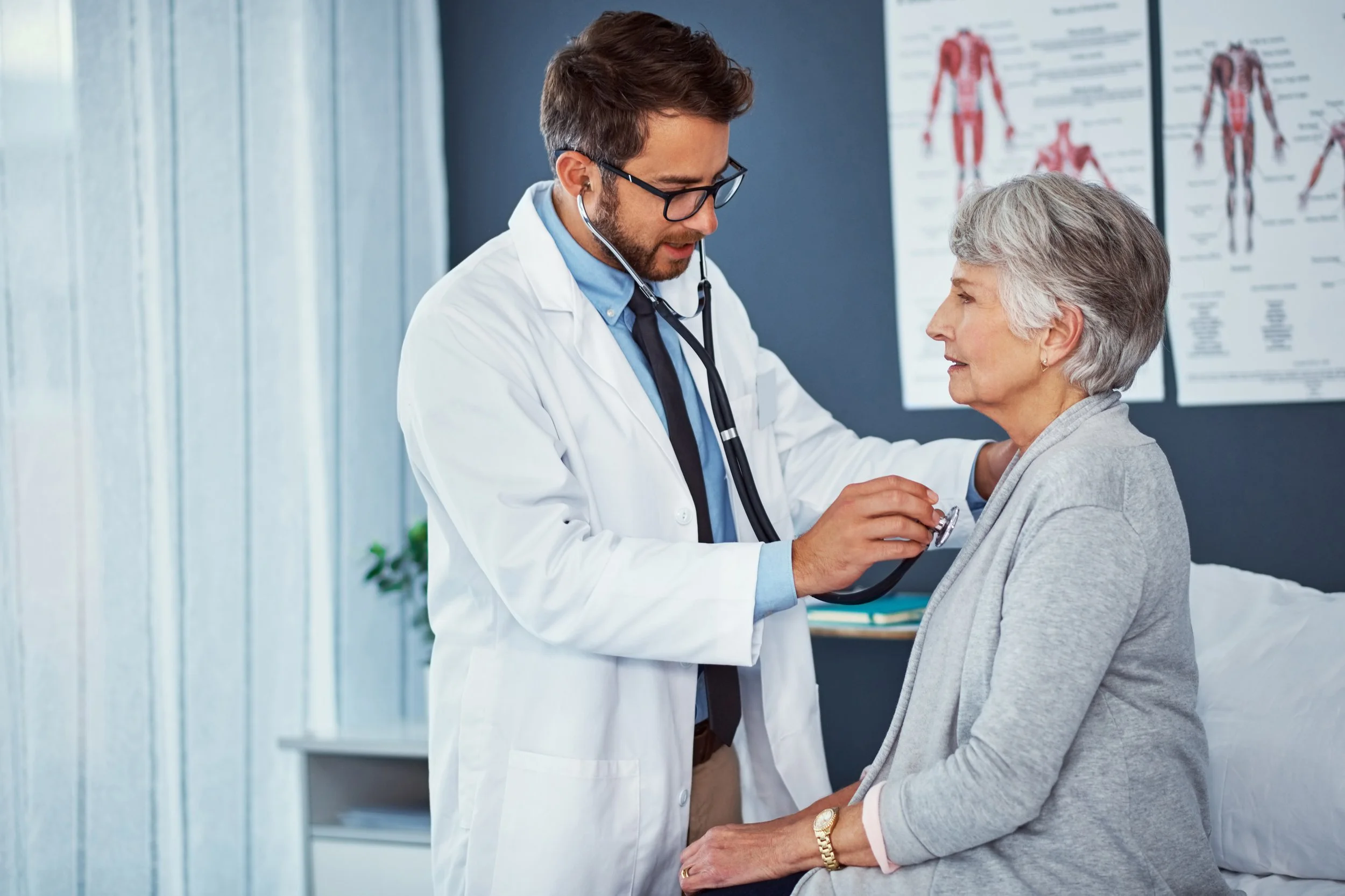 Male doctor in white coat using stethoscope on elderly woman sitting on hospital bed in exam room.