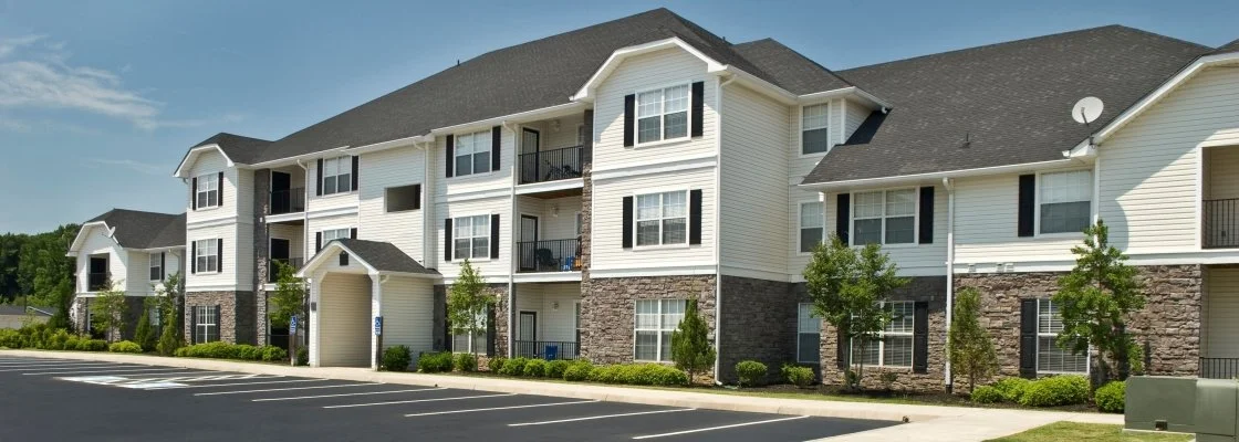 Exterior view of a three-story apartment complex with white siding, black shutters, stone accents, small trees, and a parking lot in front.