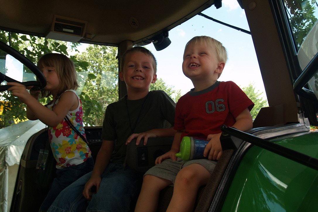 Three young children sitting inside a green tractor, smiling and laughing with trees and blue sky outside.