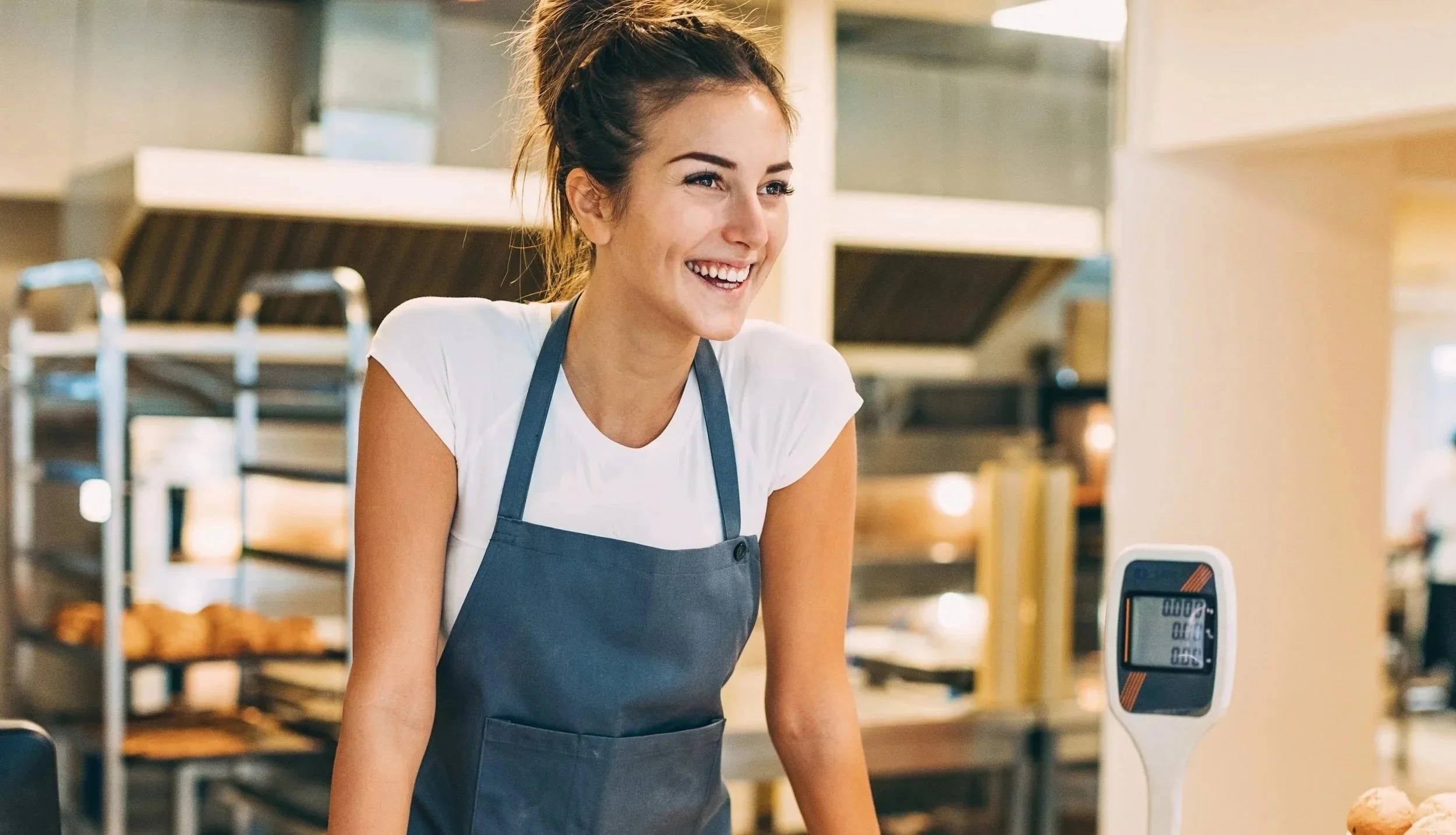 Happy woman in a bakery wearing a gray apron and white shirt, standing near a digital scale.