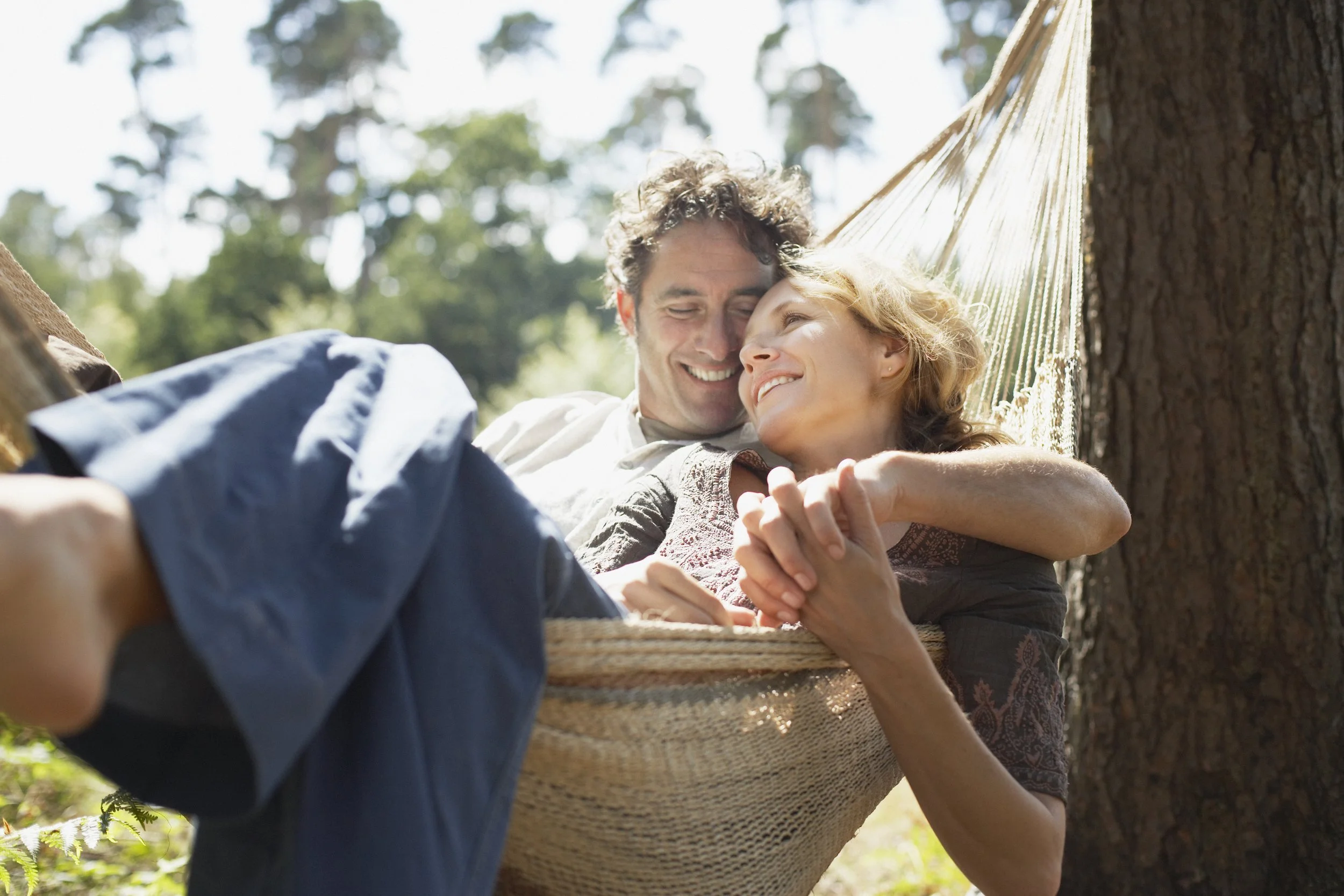 A happy couple lying together in a hammock under a tree outdoors, smiling and looking lovingly at each other.