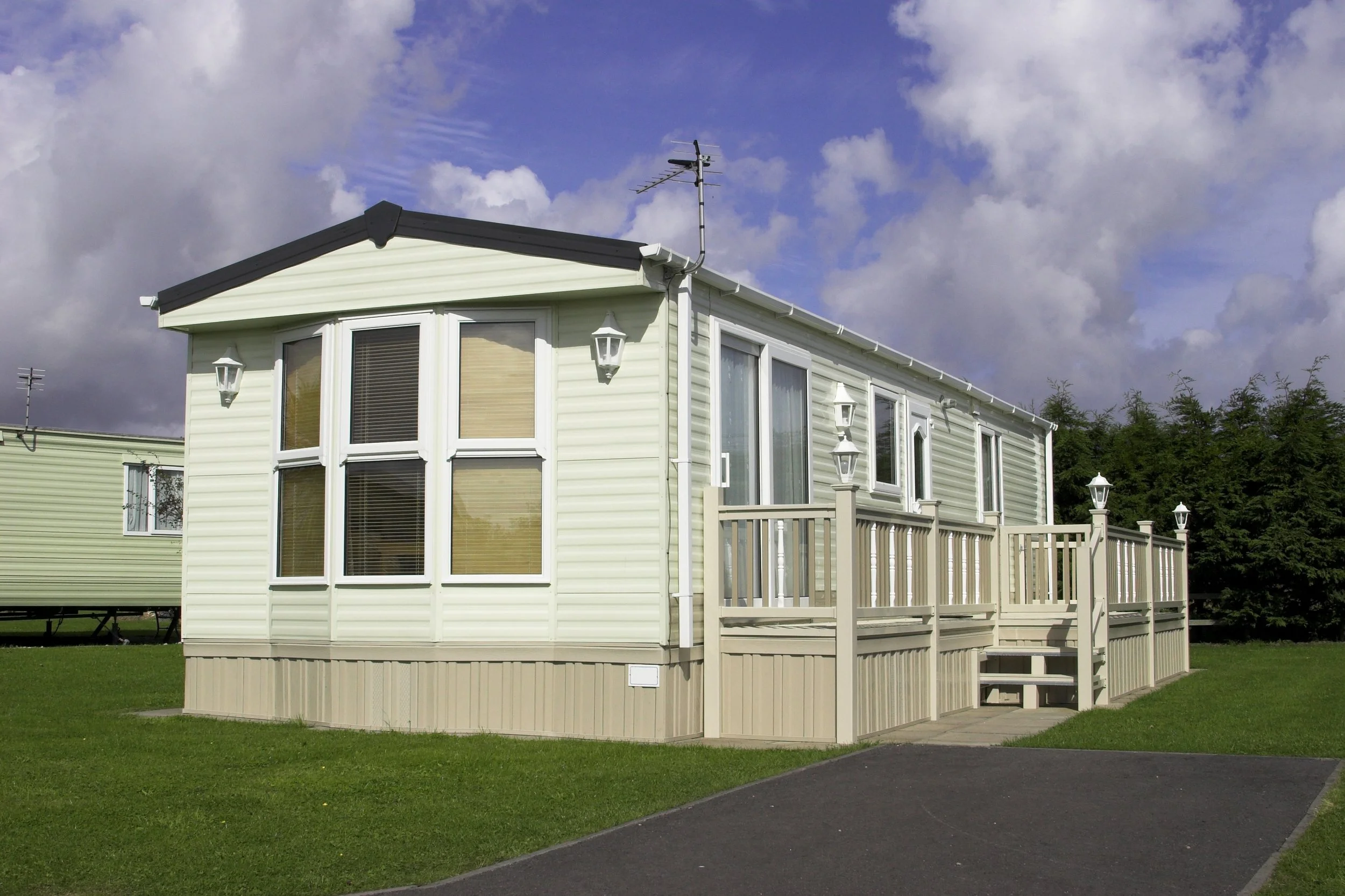 A beige mobile home with large bay windows, a small porch with a white railing and stairs, and outdoor lanterns, situated on a grassy lot with a paved driveway under a partly cloudy sky.