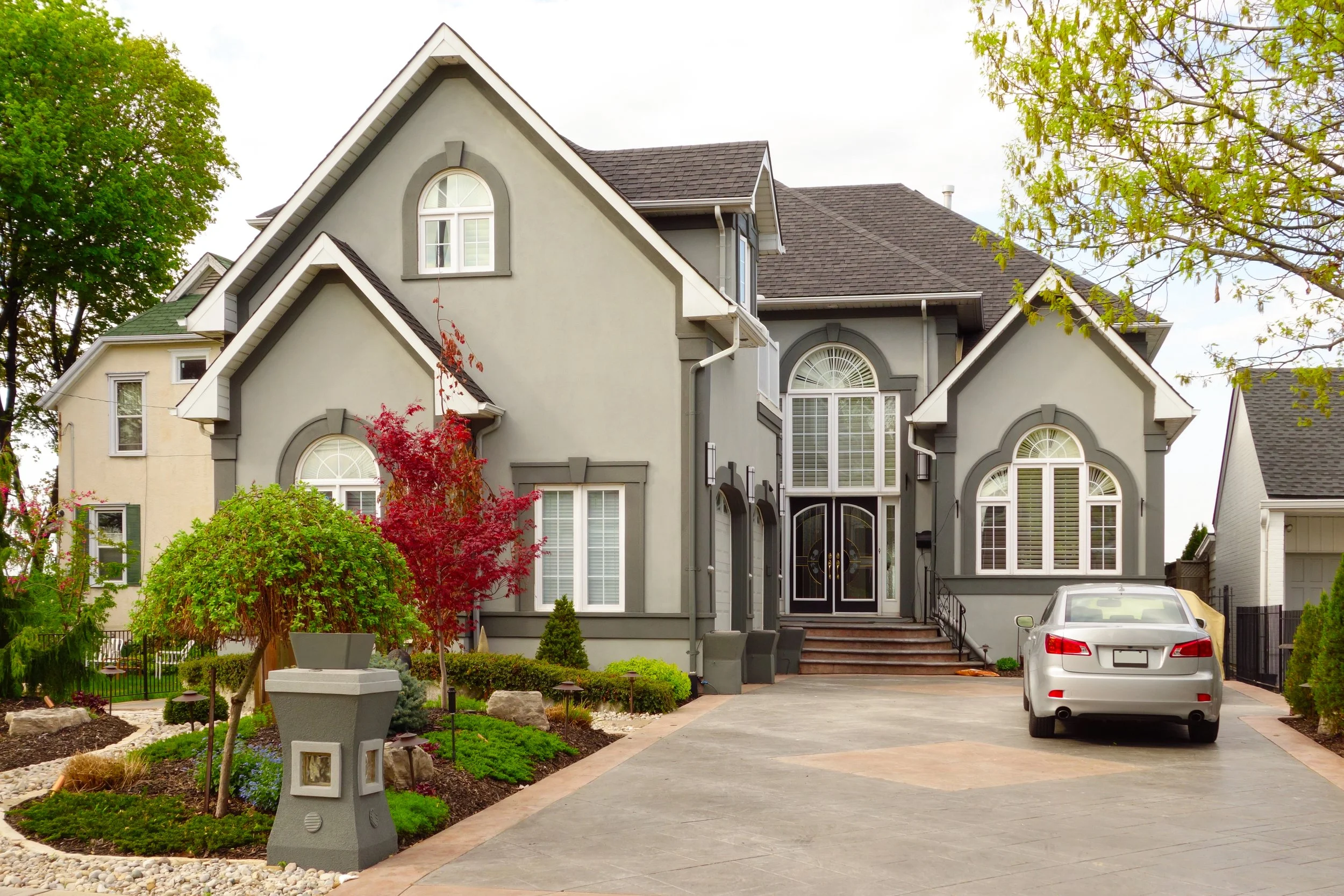 A large, gray, two-story house with white trim, front steps, and a driveway with a white car parked. The house features multiple windows with decorative arch designs and has landscaped greenery and trees in the front yard.