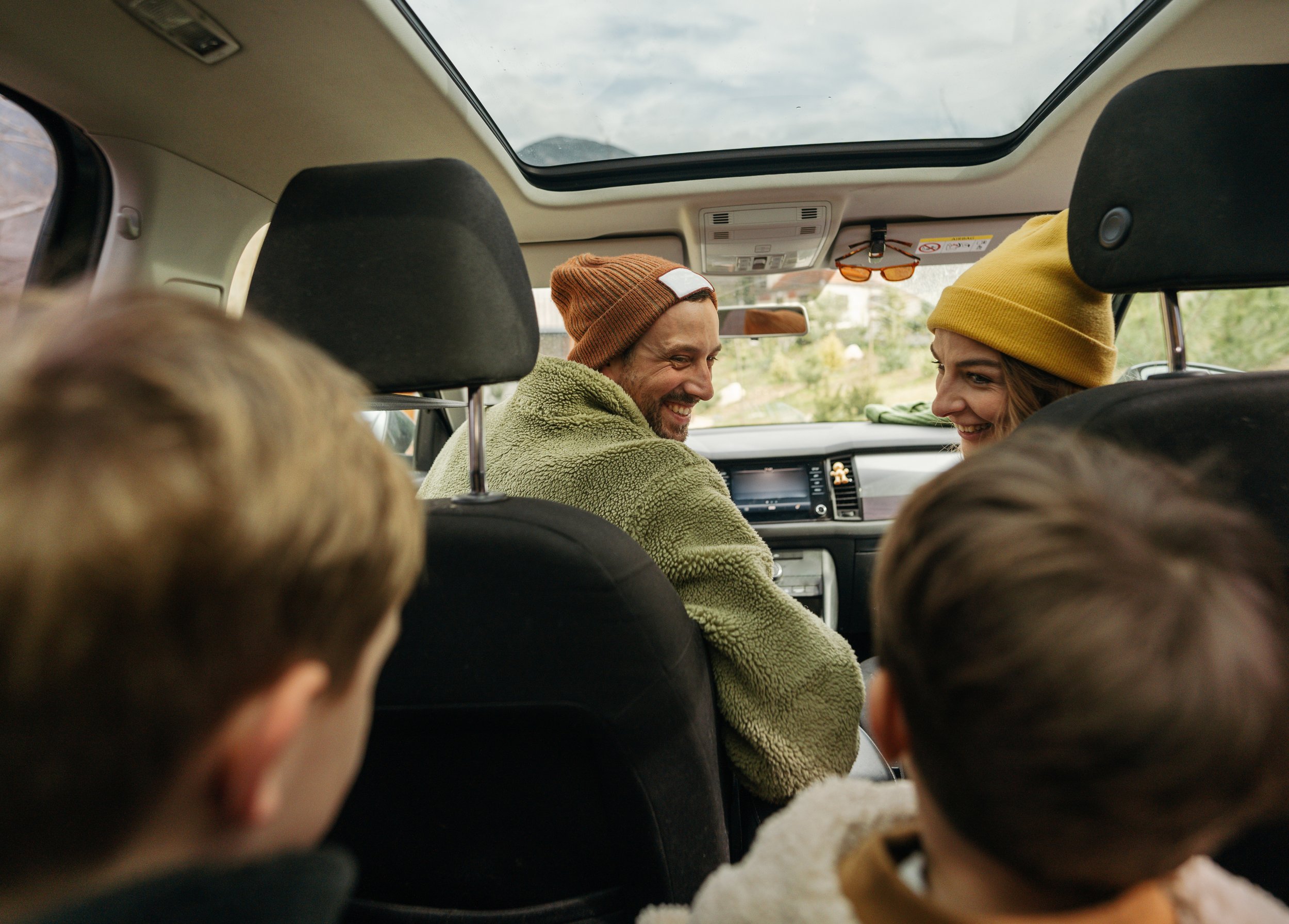 A family inside a car, with two adults smiling at each other in the front seats, and two children visible from behind in the back seats, during daytime with a scenic landscape outside.