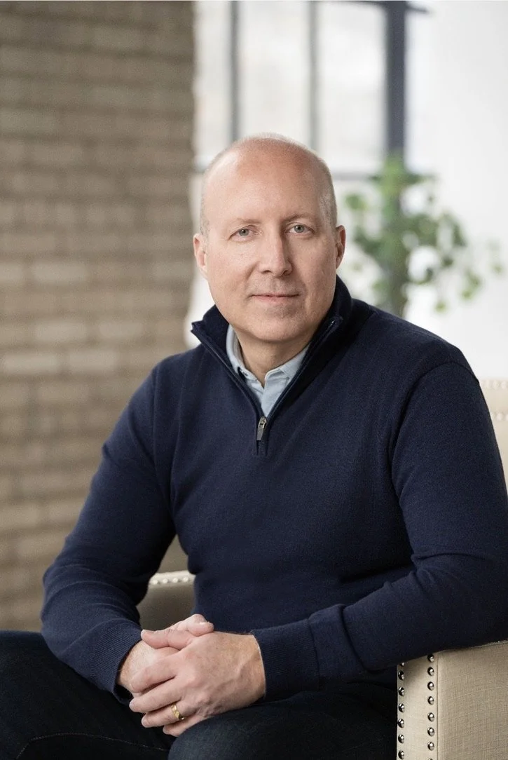 A man with a bald head wearing a navy blue zip-up sweater over a collared shirt, sitting indoors with a neutral background and a green plant in the background.