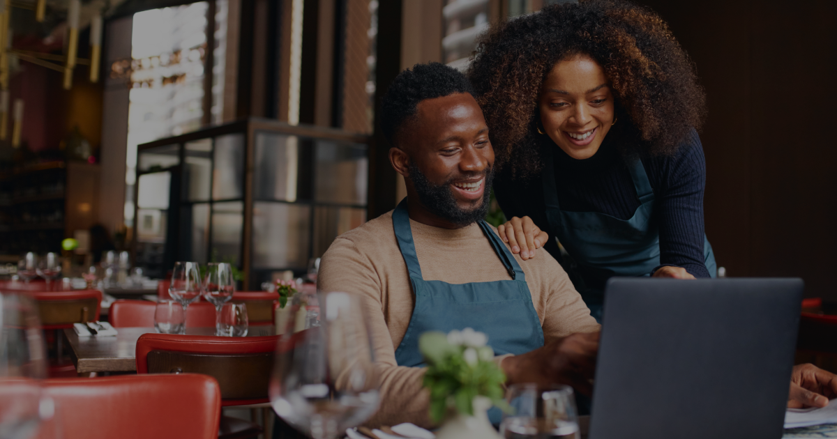 A man and a woman in a restaurant looking at a laptop, smiling, with tables set for dining in the background.
