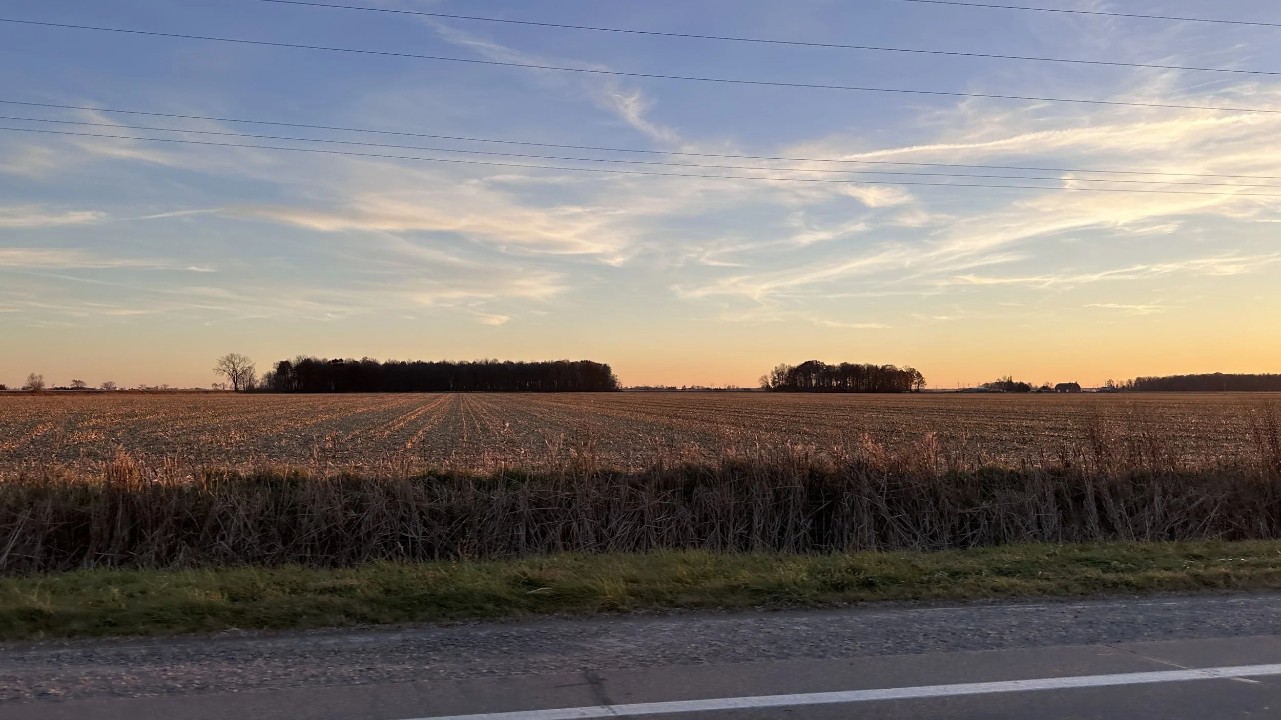 A rural landscape at sunset with a wide open field, trees in the background, and power lines above. The foreground shows a gravel road and a grassy roadside.