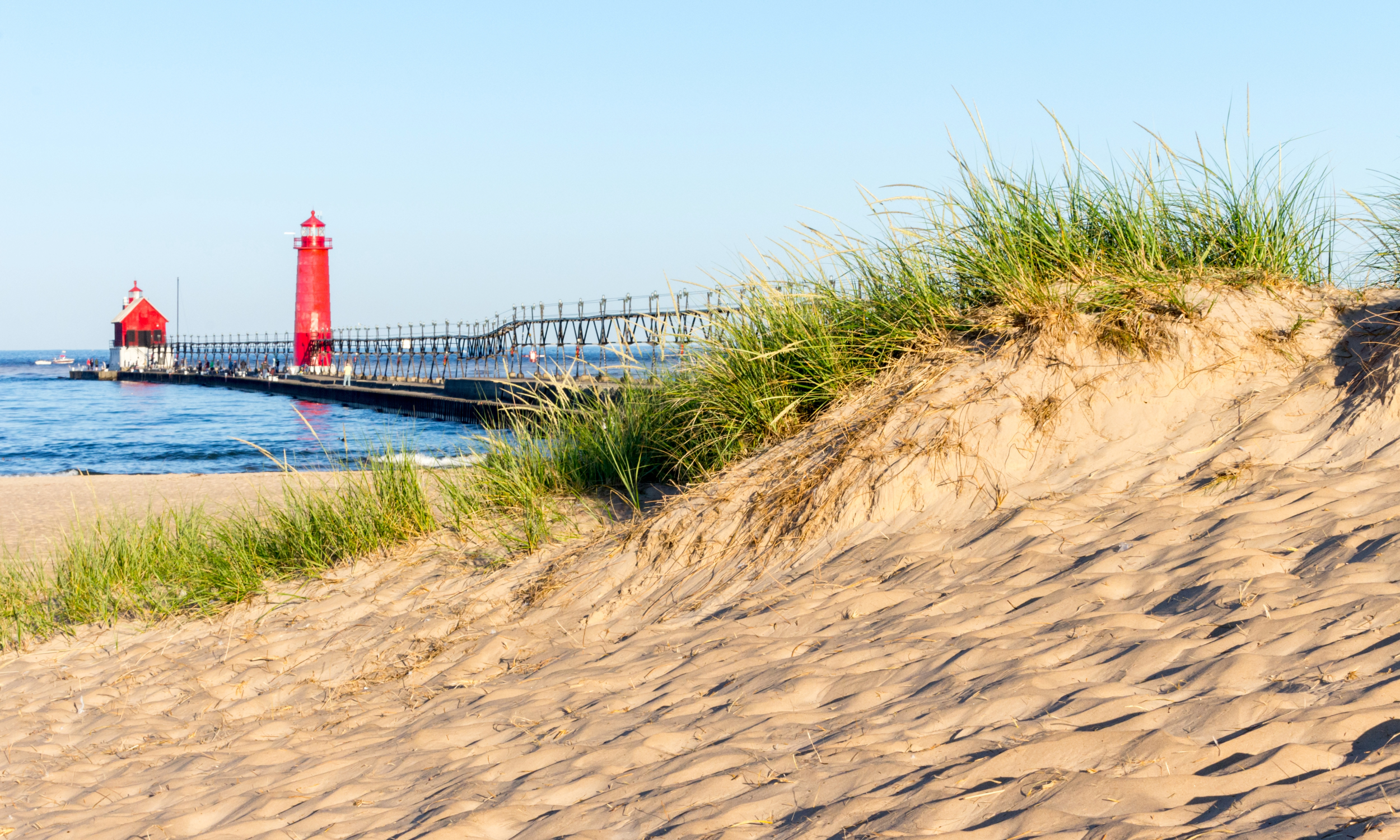Sandy dunes with grassy plants in the foreground and a red lighthouse on a pier extending into the water in the background on a clear day.