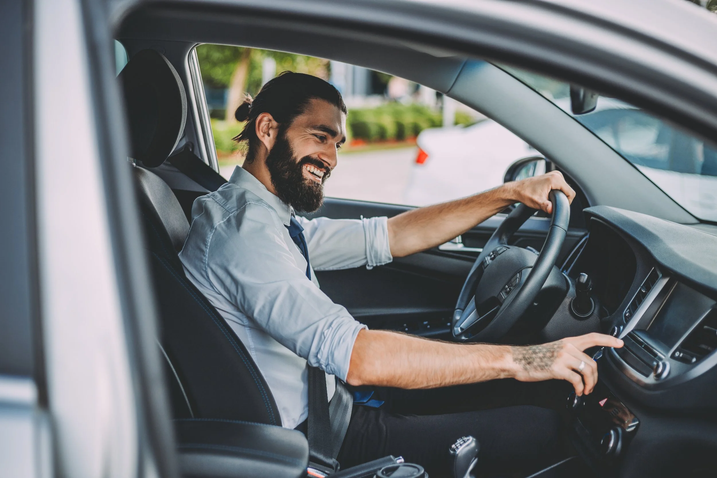 A man with a beard and long hair tied back, smiling while sitting in the driver's seat of a vehicle, reaching for the dashboard.