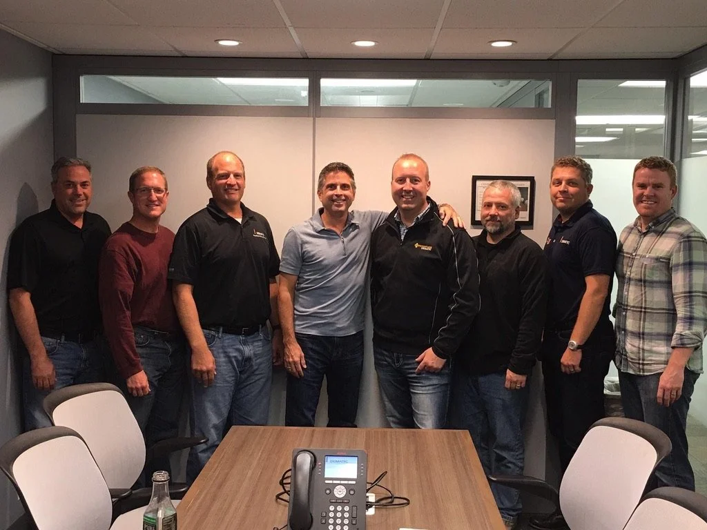 Group of nine men standing in a conference room, smiling, with a conference table and phone in the foreground.