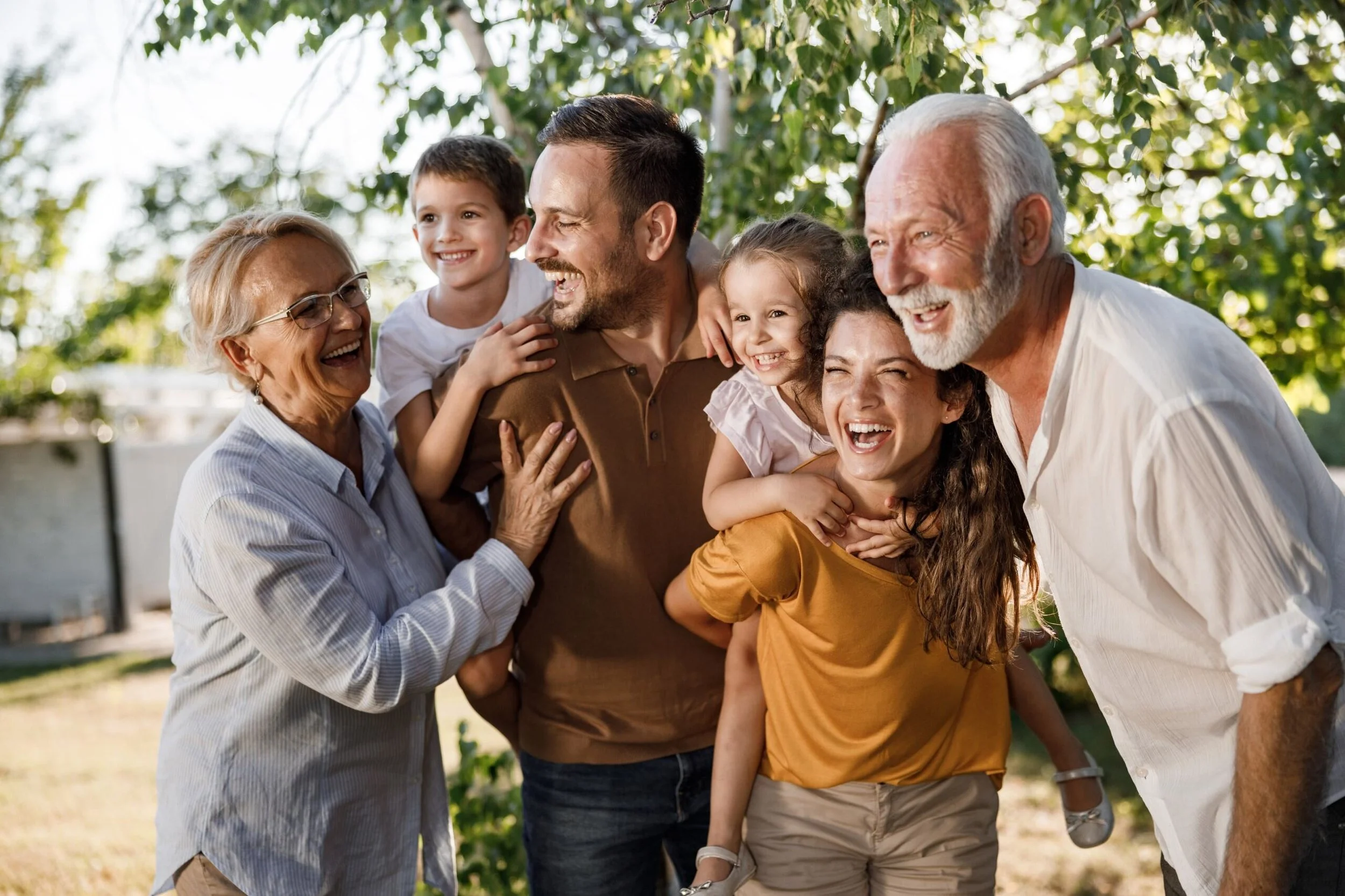 A multi-generational family of six enjoying a happy moment outdoors, laughing and hugging in a park-like setting with trees.