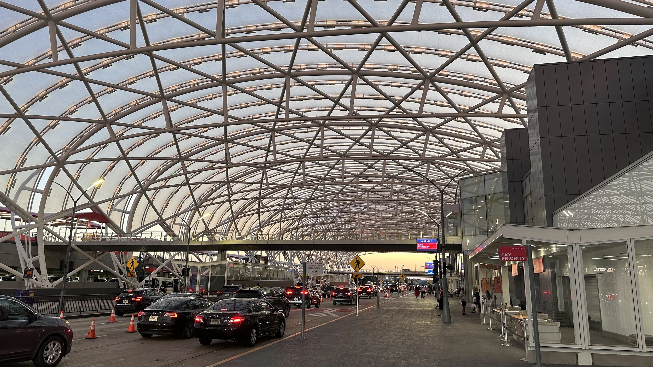 Modern airport terminal with a large, curved glass and steel roof structure. Vehicles are parked or moving near the terminal, and people are walking or waiting. Signage indicates Sky Priority service.