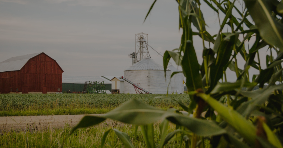 Farmland with a red barn, metal grain silo, and agricultural equipment, framed by green crops in the foreground and overcast sky.