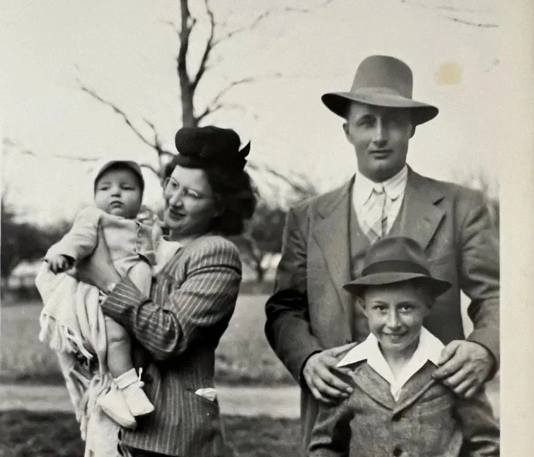 Black-and-white photo of a family outdoors, with a woman holding a small child and a man with a young boy standing beside him, trees in the background.