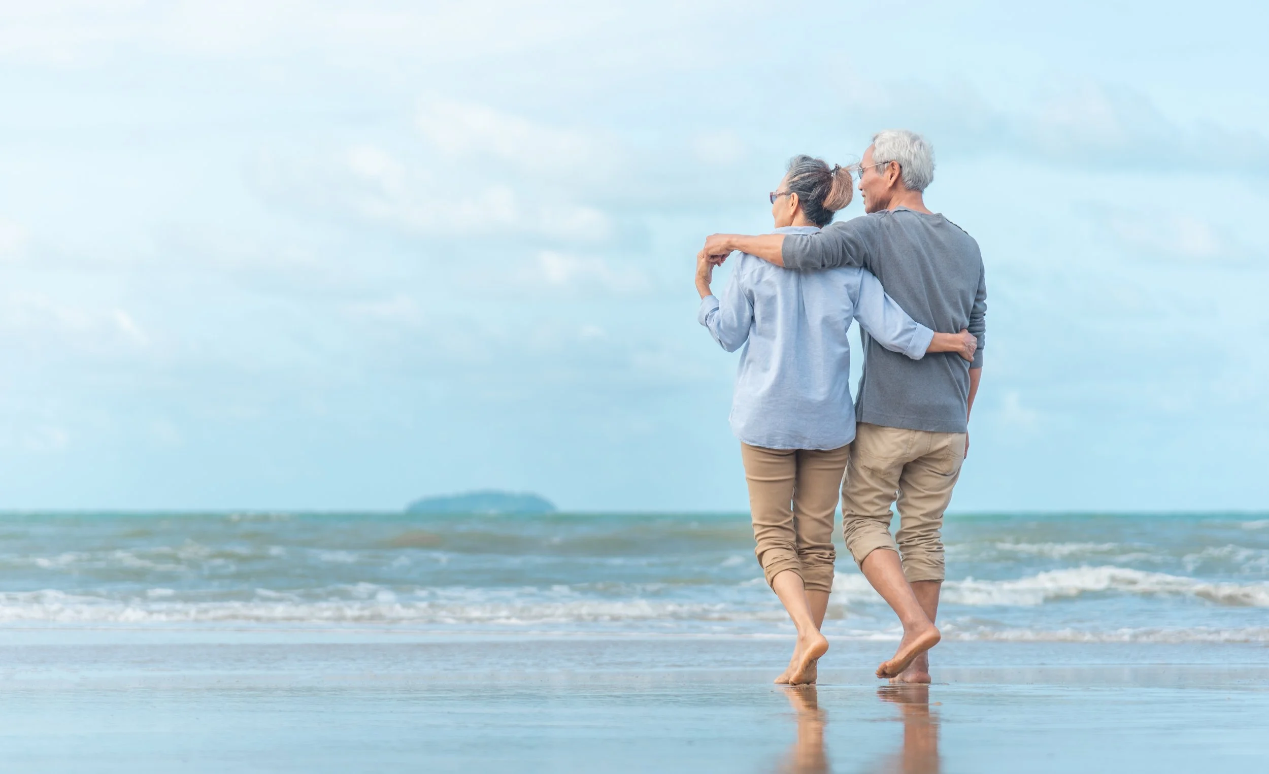 An elderly couple walking barefoot on the beach, embracing and looking out at the sea under a partly cloudy sky.