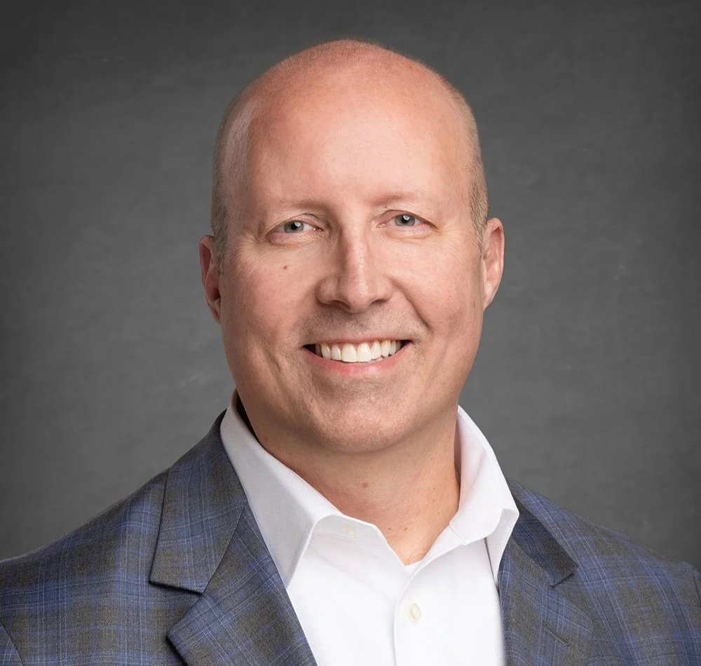 Headshot of a smiling man with a bald head, wearing a white shirt and a dark checked blazer, against a gray background.