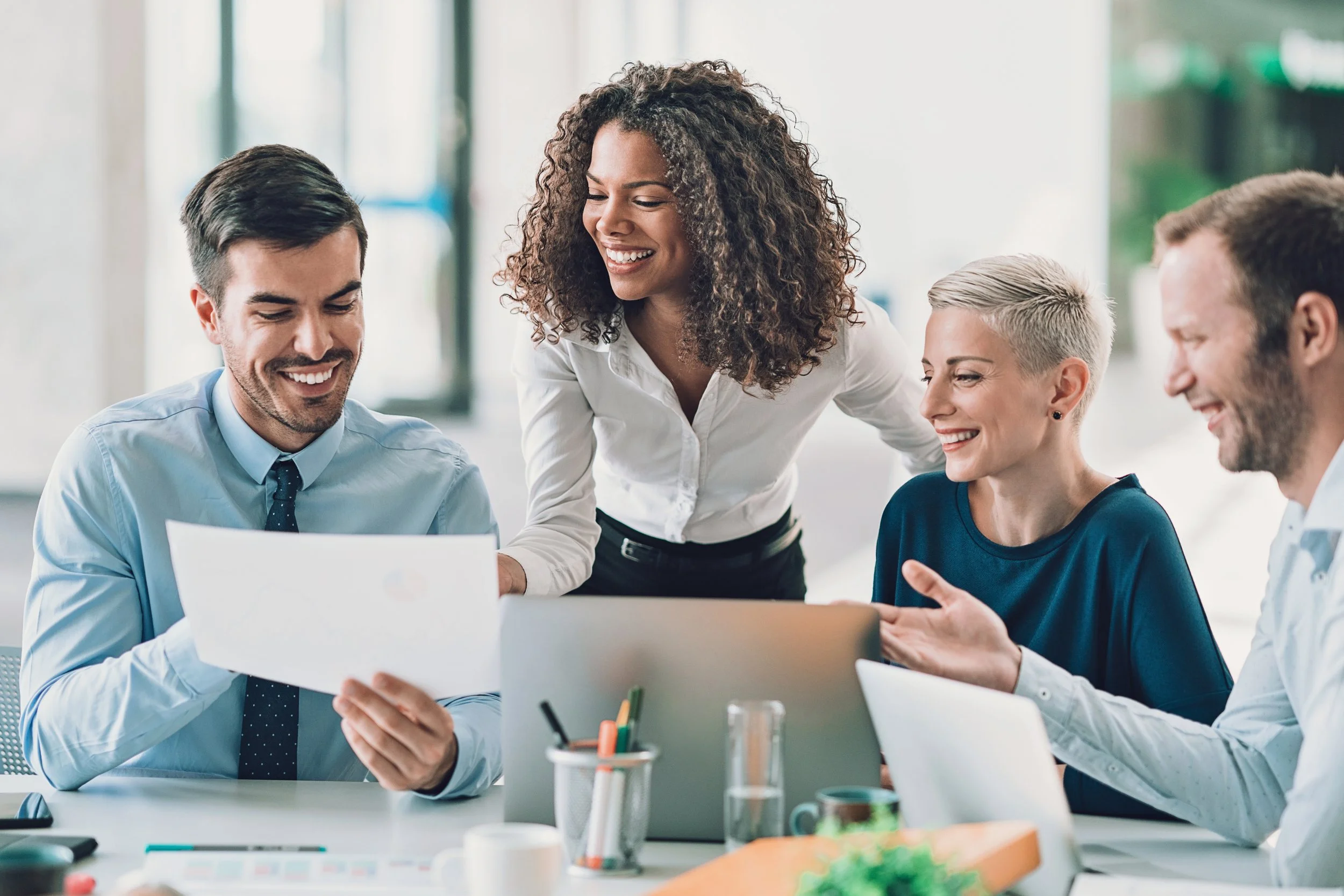 Four diverse colleagues smiling and discussing work in an office, with laptops, documents, and office supplies on the table.