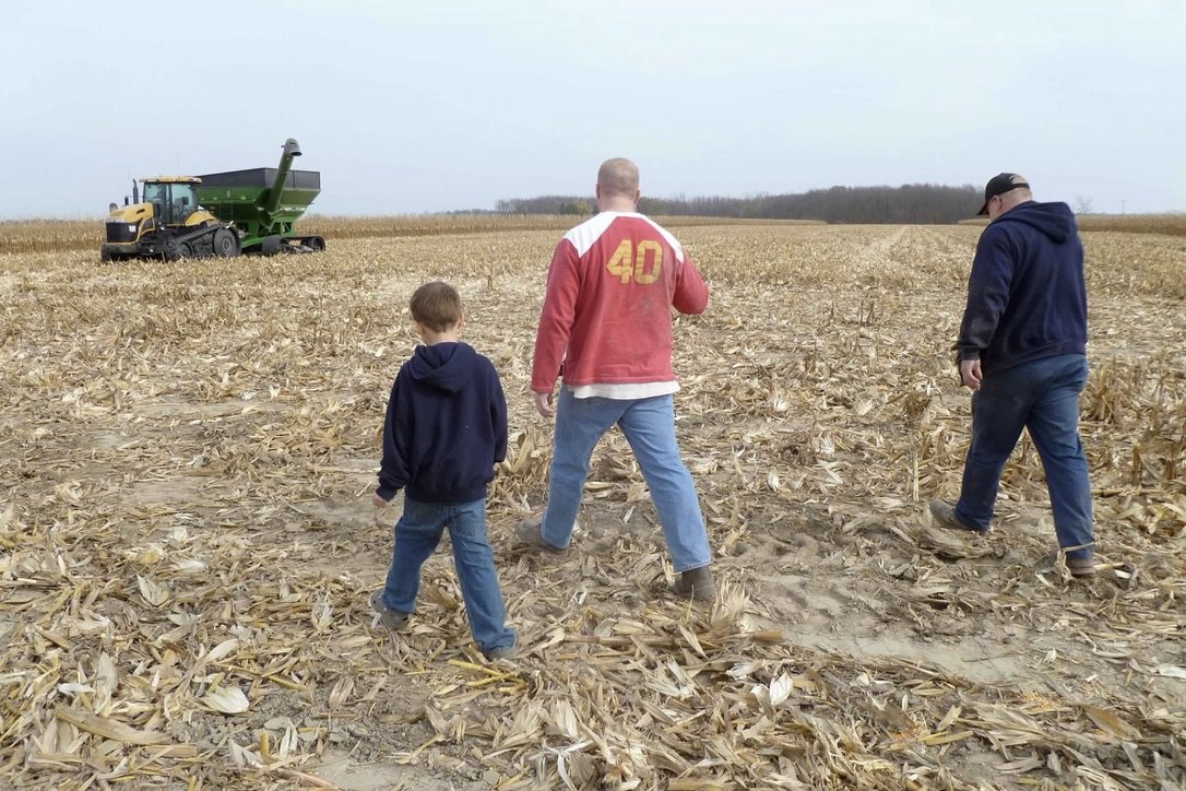 Three people, two adults and a child, walk through a harvested cornfield with a tractor in the background under an overcast sky.
