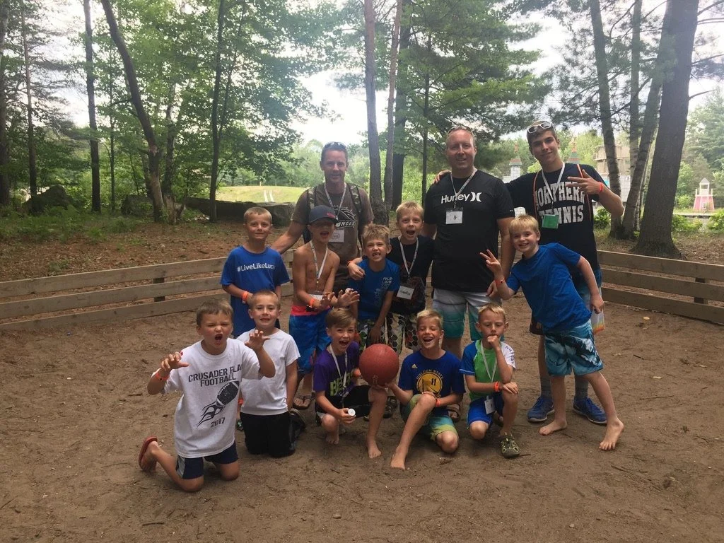 Group of kids and adults playing basketball outdoors in a wooded area, some kids wearing medals.