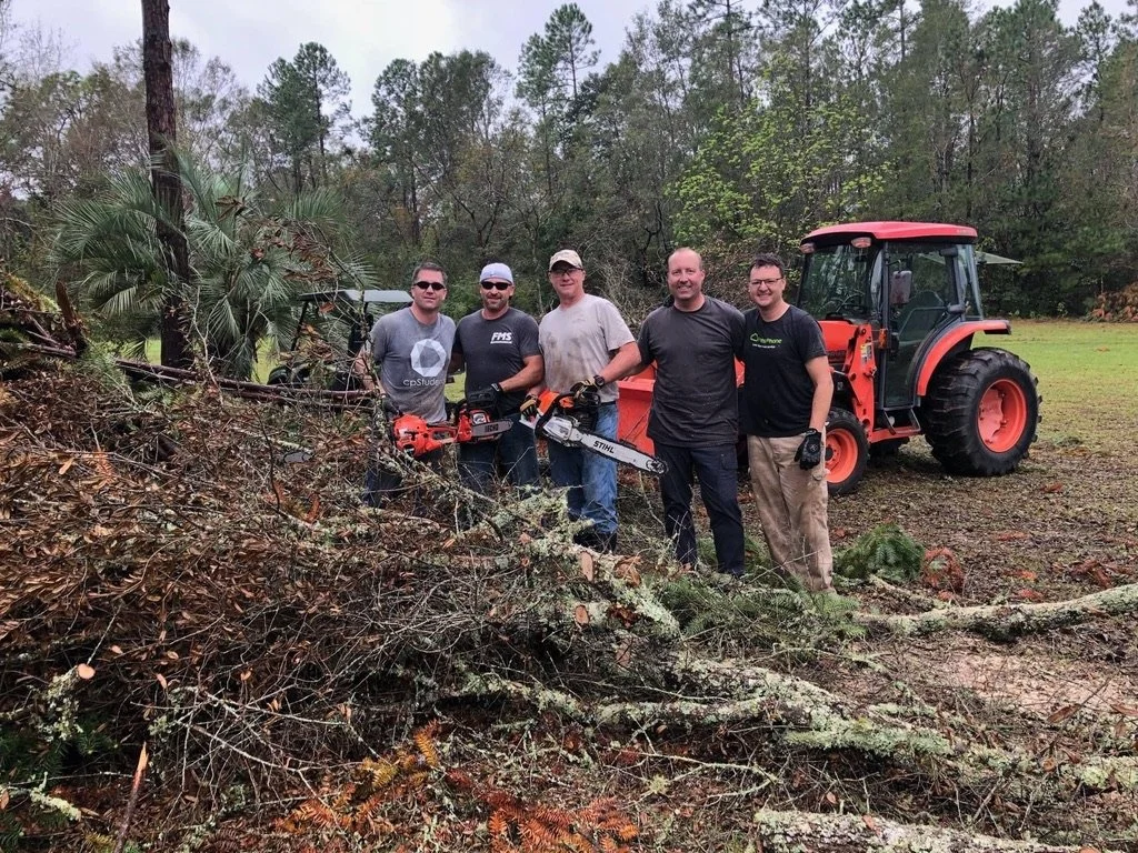 Five men standing outdoors in front of a recently cut fallen tree and a red tractor, holding chainsaws, in a wooded area.