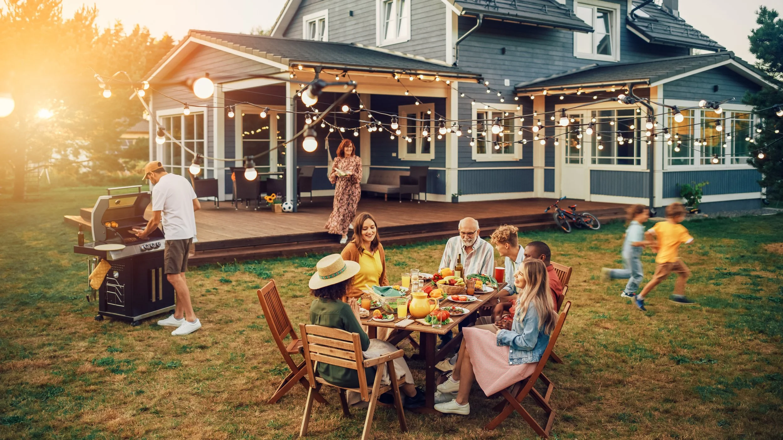 Family gathering on a backyard patio with string lights, a man grilling, children playing on the grass, and friends sitting at a table enjoying food and drinks.