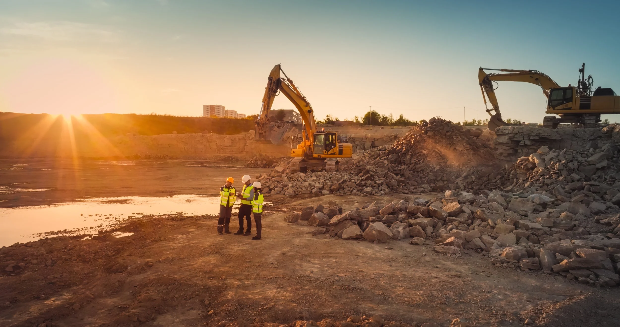 Construction site at sunset with two excavators demolishing a structure and three workers in safety vests and helmets discussing in the foreground.