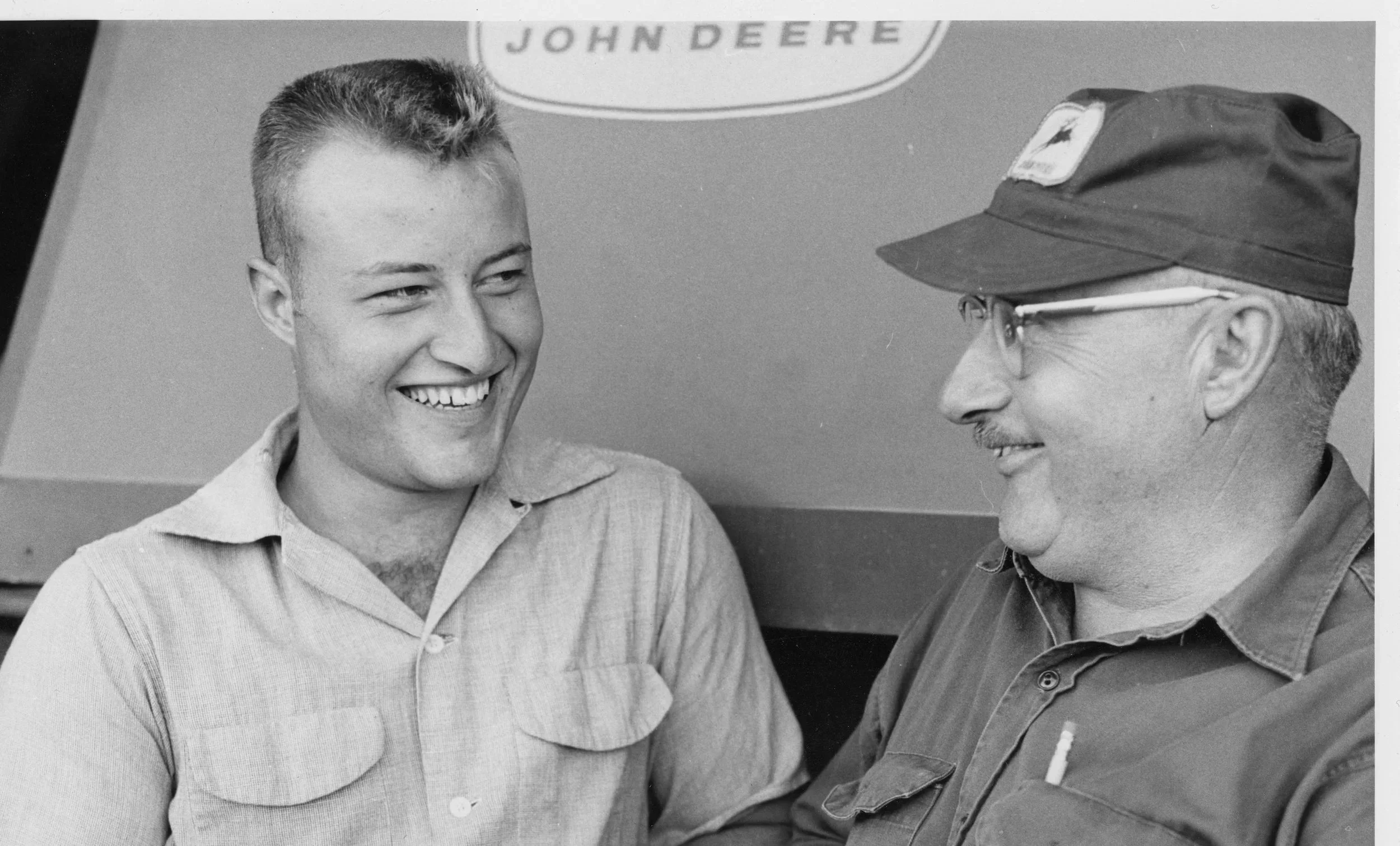 Black and white photograph of a young man and an older man smiling at each other. The young man has short hair and is wearing a collared shirt. The older man is wearing glasses and a cap with an emblem, and a shirt with a pen in the pocket. There is a partial visible sign above them that reads "JOHN DEERE."