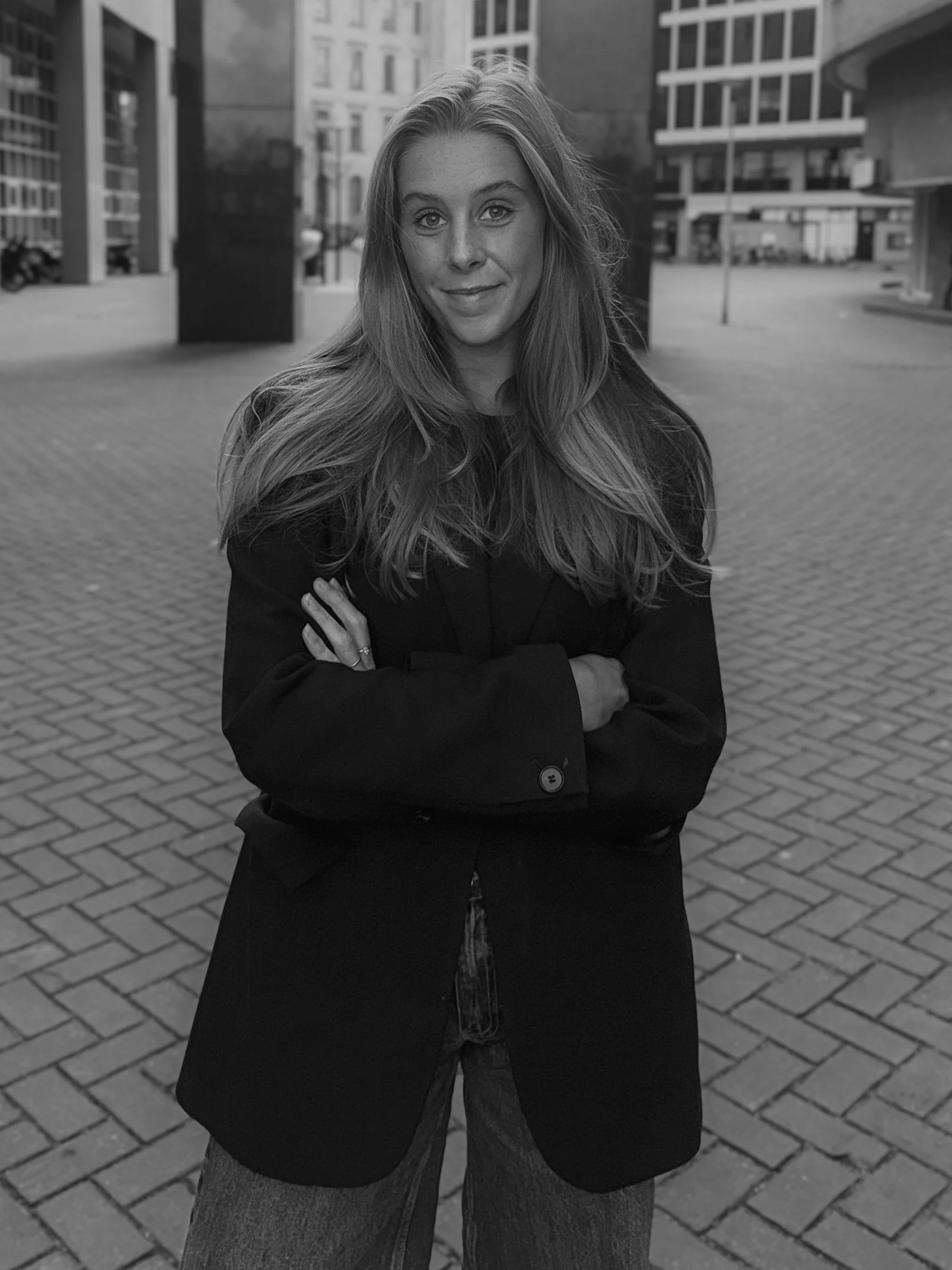 A woman with long hair crossing her arms, standing on a city sidewalk with buildings in the background.