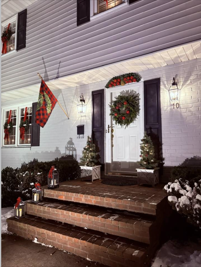Festively decorated house entrance with Christmas wreath on the door, small illuminated Christmas trees, lanterns, and snow on the ground and bushes.