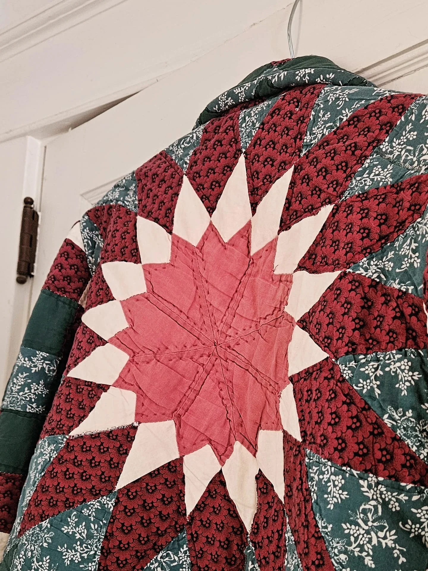 Close-up of a quilt featuring a large red and white star pattern with green and red floral fabric surrounding it, hanging on a white wall.