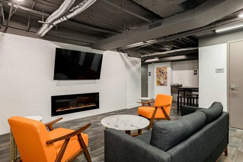 Modern lounge with a flat-screen TV, fireplace, orange armchairs, a gray sofa, a round marble coffee table, and a painting in a basement setting.