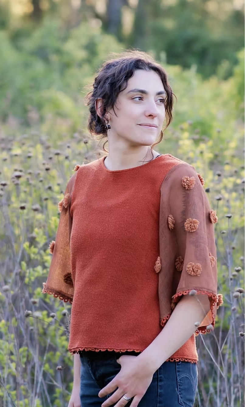 A woman with short, curly dark brown hair and earrings standing outdoors in a field of tall grass and plants, wearing a burnt orange top with sheer, decorated sleeves, looking to her left with a smile.