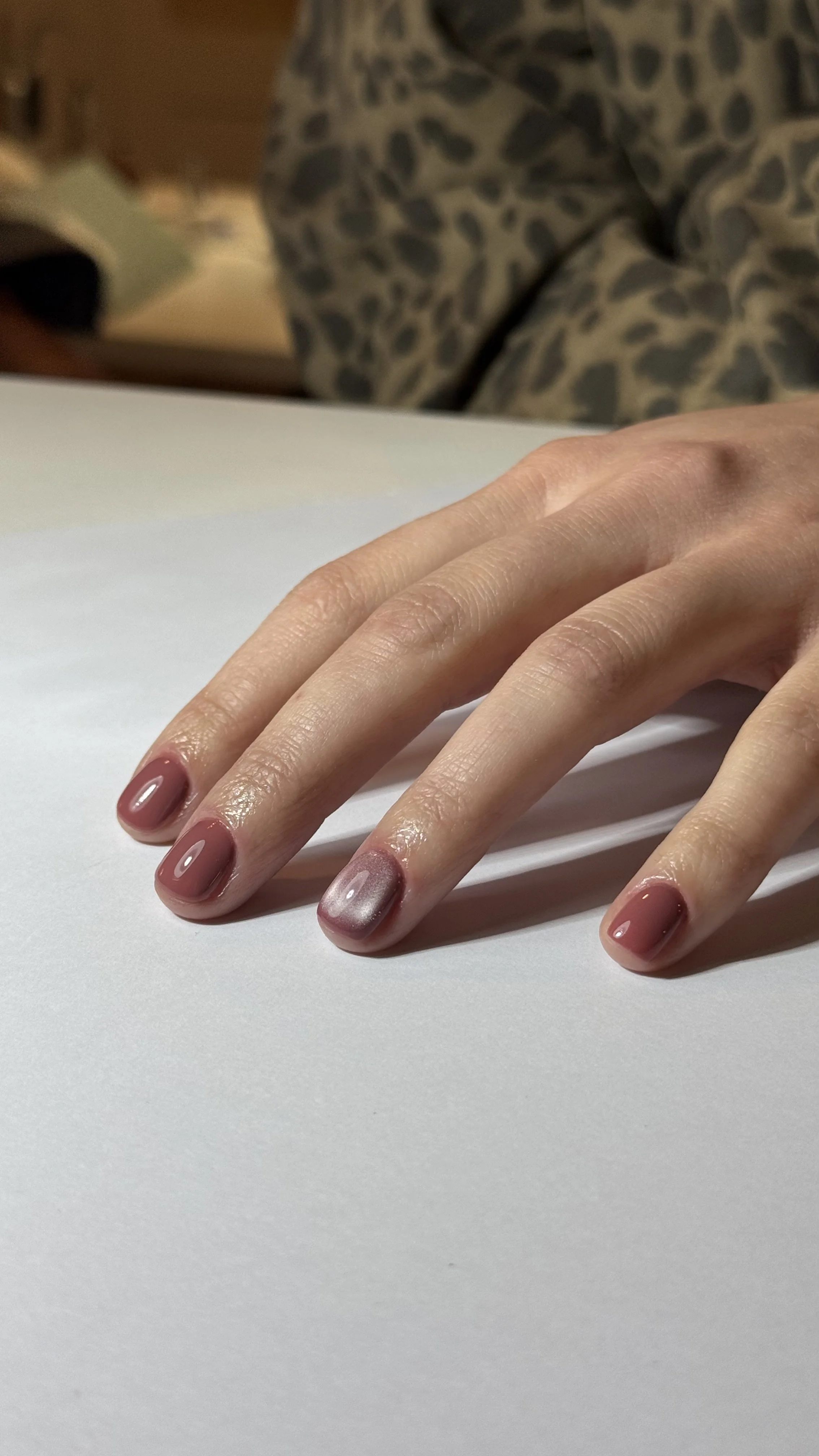 Main avec des ongles vernis en nuances de rose et de gris, posé sur une surface blanche.