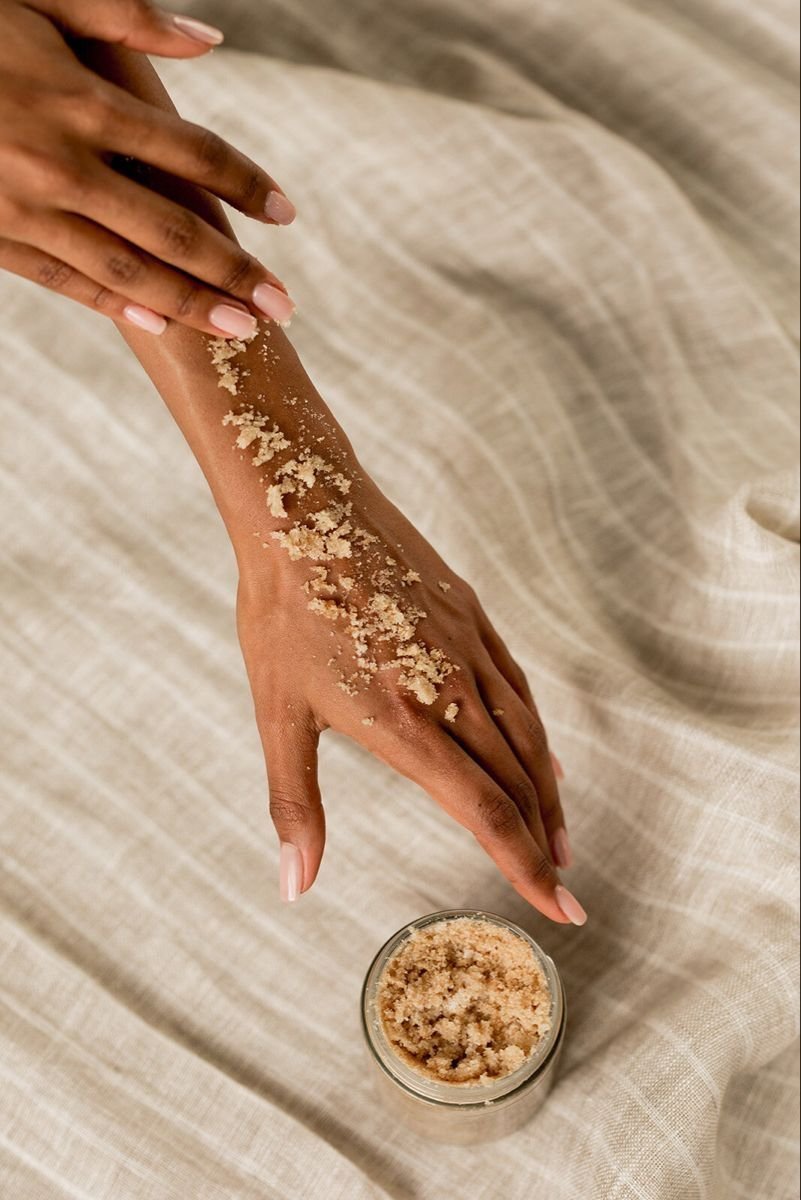 Une main habile appliquant un exfoliant au sucre sur l'avant-bras d'une autre personne. Le fond est une surface en tissu beige.