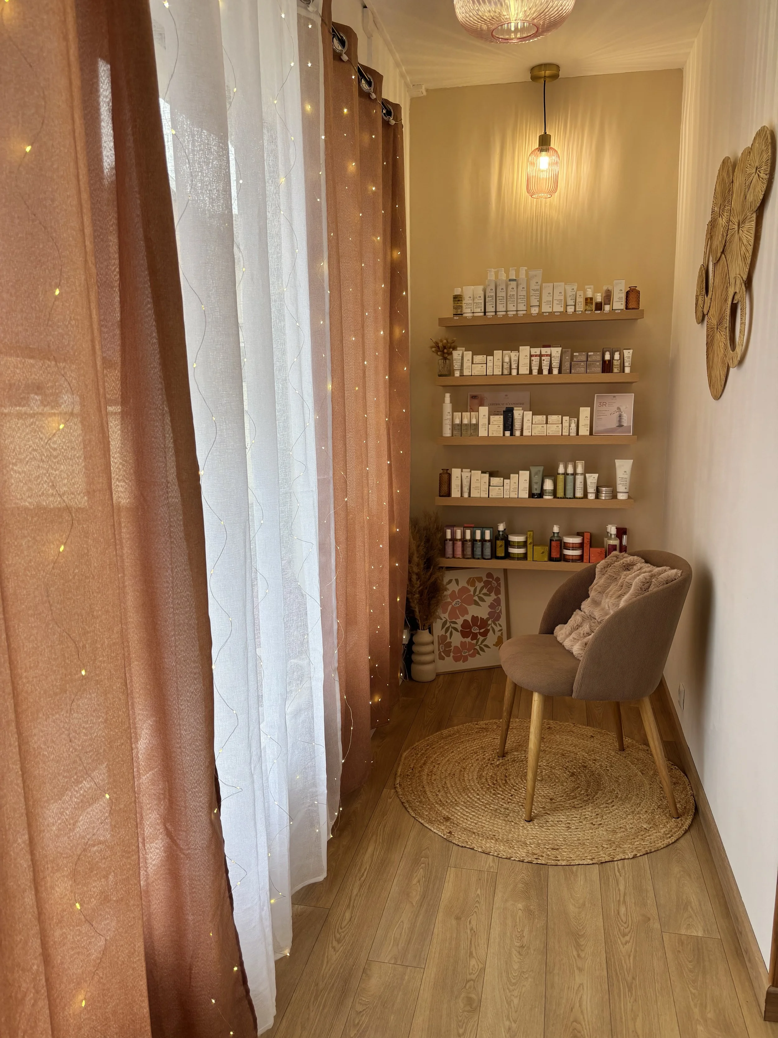 Un institut de beauté  avec un fauteuil en tissu beige, un tapis rond en fibre naturelle, des étagères en bois avec des produits cosmétiques, une lumière suspendue douce, des rideaux colorés avec guirlande lumineuse.