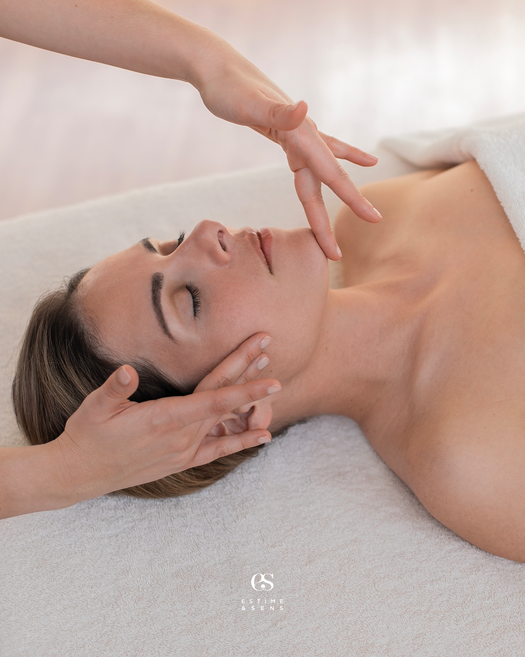 Une femme reçoit un massage visage dans un institut, allongée sur une table, avec un drap blanc partiel couvrant son corps.
