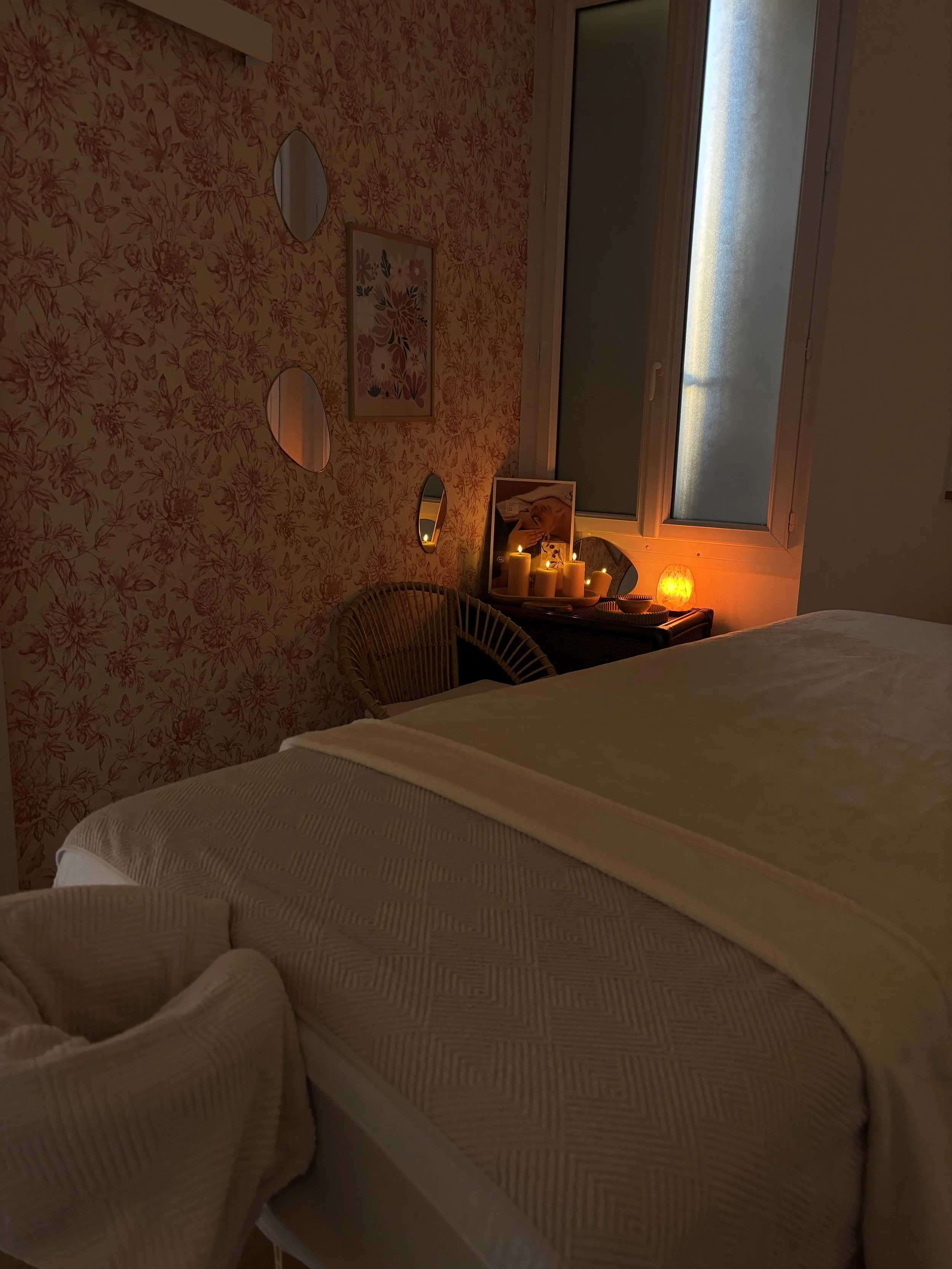 Une chambre douce et chaleureuse avec un lit fait, une fenêtre avec volets, un mur tapissé de motifs floraux, et une table de chevet avec des bougies, un miroir, un cadre photo et une lampe orange