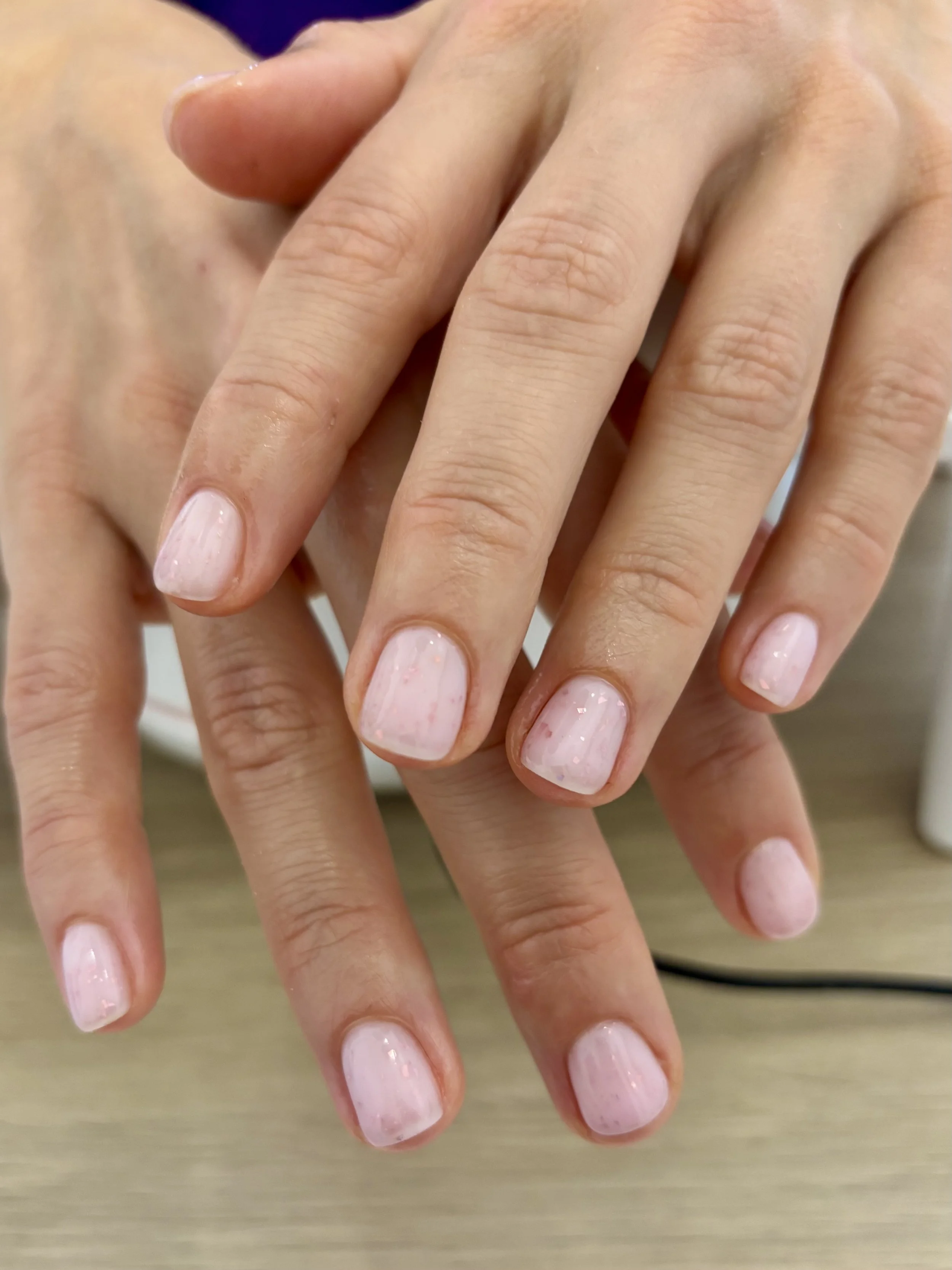 Une paire de mains avec des ongles vernis en rose clair, posées l'une sur l'autre sur une surface en bois, avec des ongles courts et bien entretenus.
