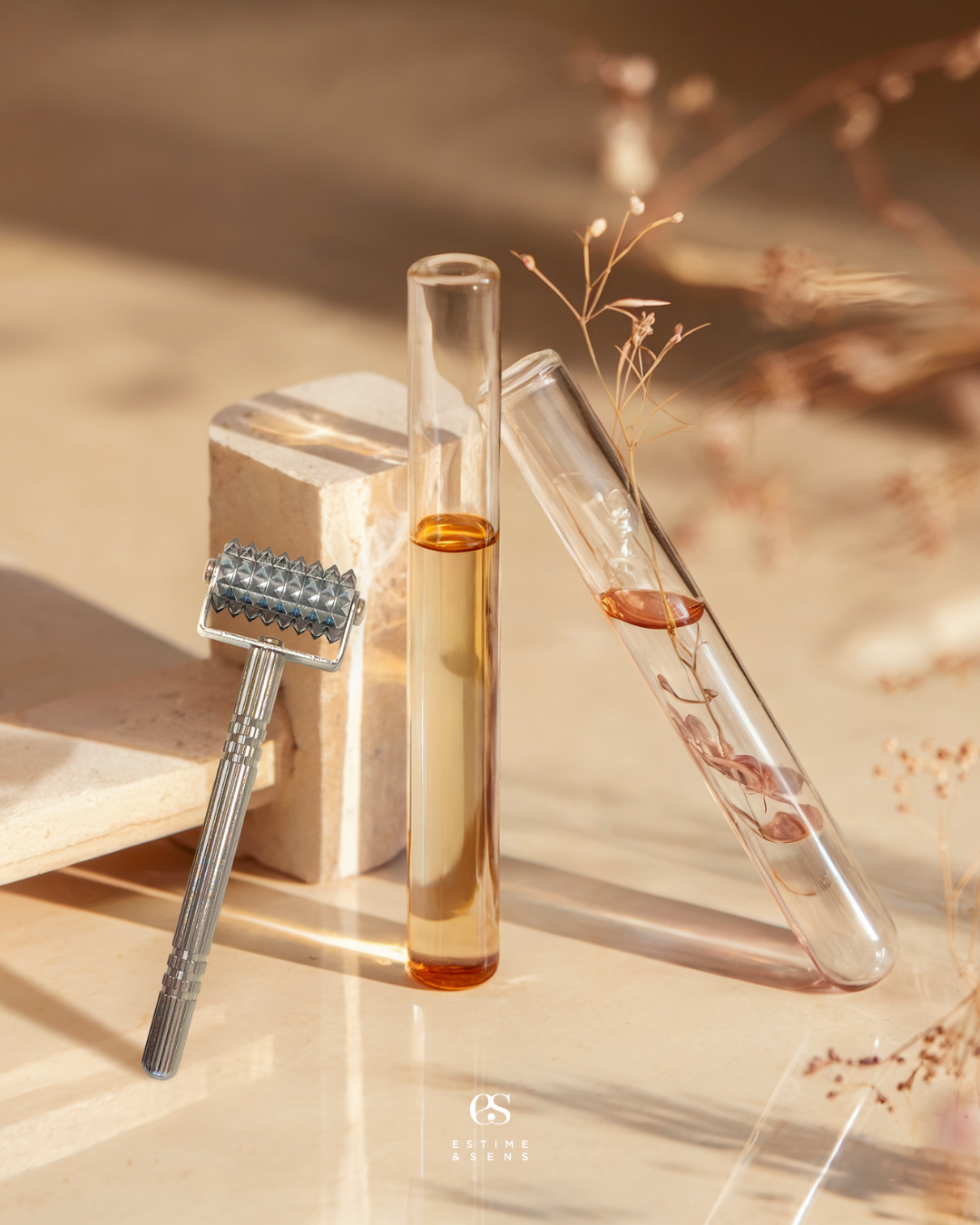 Deux tubes à essai en verre, l'un rempli d'un liquide orange clair, l'autre avec un liquide légèrement rosé, posés sur une surface beige avec un petit pilon en métal à côté et un bloc en pierre derrière, avec des petites branches sèches en décoration.