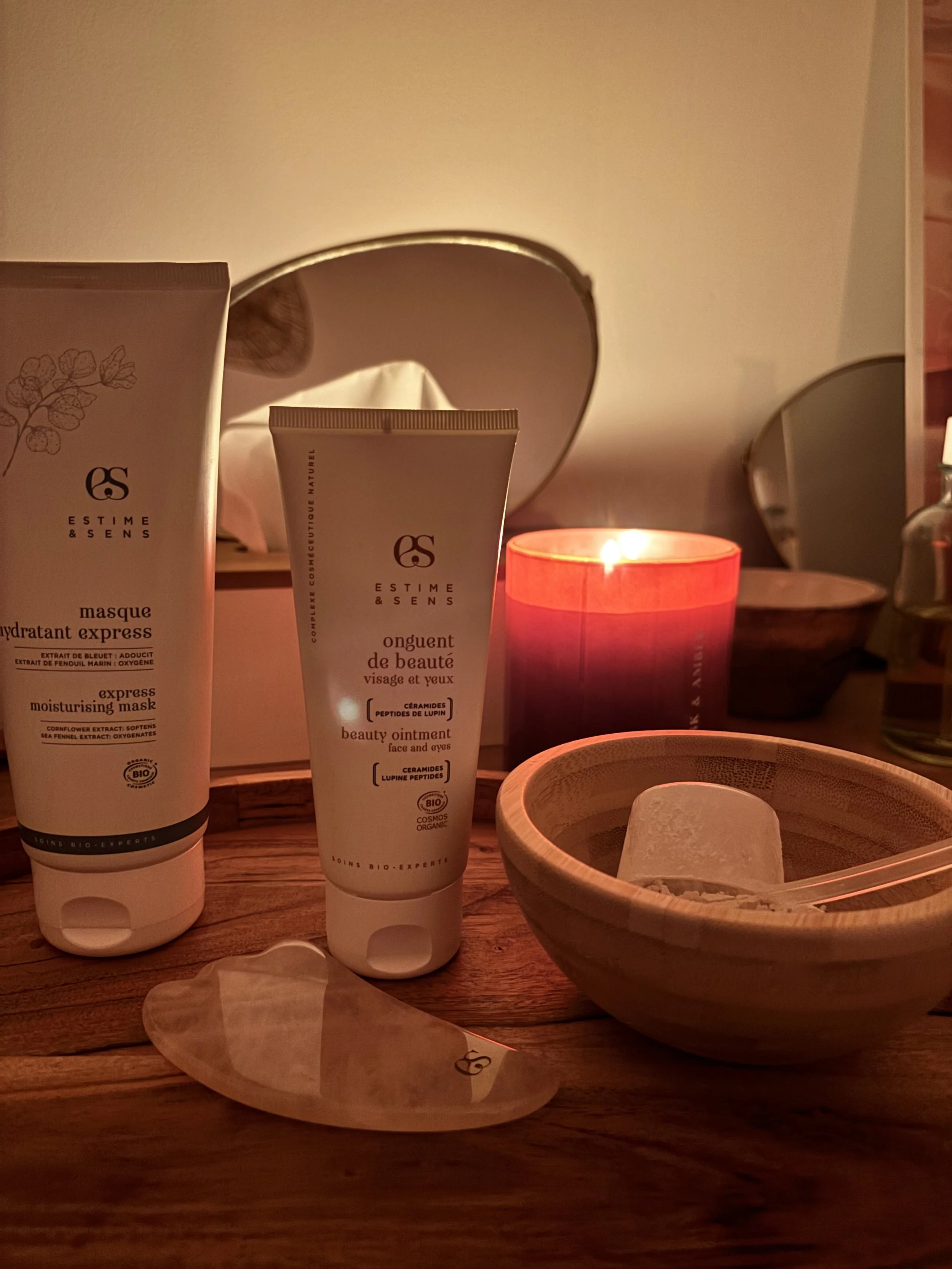 Des produits cosmétiques bio sur une table en bois, avec une bougie allumée dans un verre rouge, un bol en bois avec des pierres ou esthétiques, et un miroir en arrière-plan.