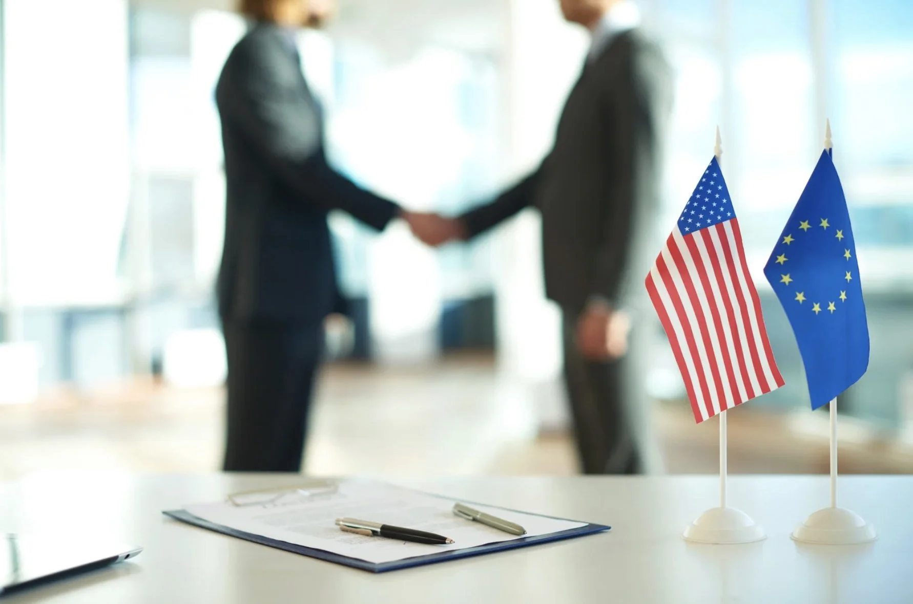 Duas pessoas apertando as mãos ao fundo, ambas usando terno. Na mesa à frente, há duas miniaturas de bandeiras dos Estados Unidos e da União Europeia, além de uma pasta com documentos e duas canetas.