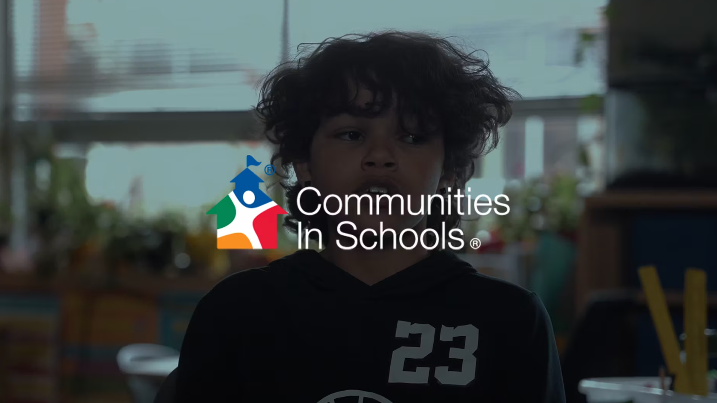A young boy with the number 23 on his sweatshirt sits in a classroom in front of a window, with the logo of Communities In Schools overlaid.