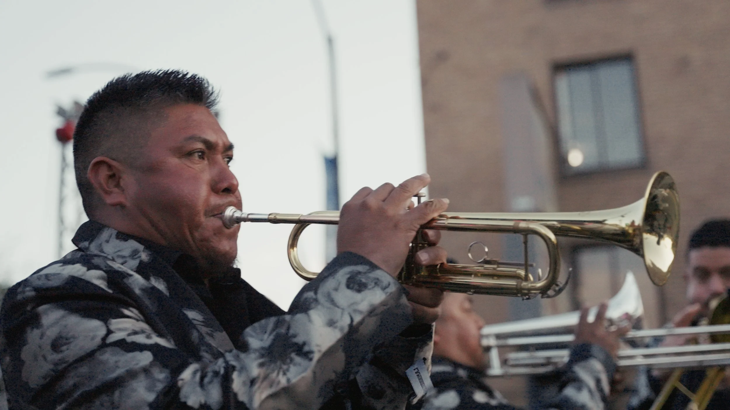 A man playing trumpet outdoors, with other musicians in the background, during a performance or parade.