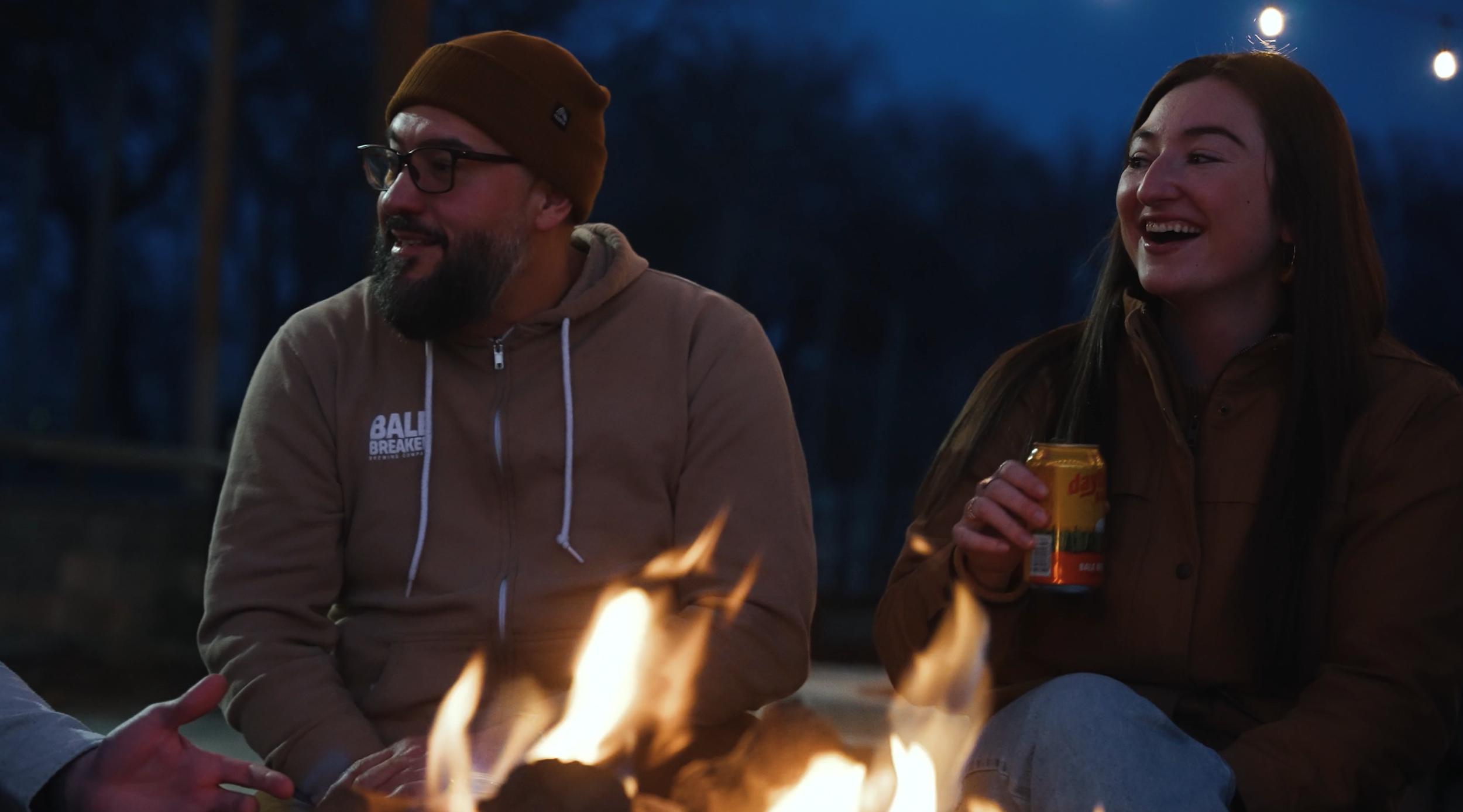 Two people sitting around a campfire at dusk, smiling and enjoying drinks, outdoors with string lights overhead.