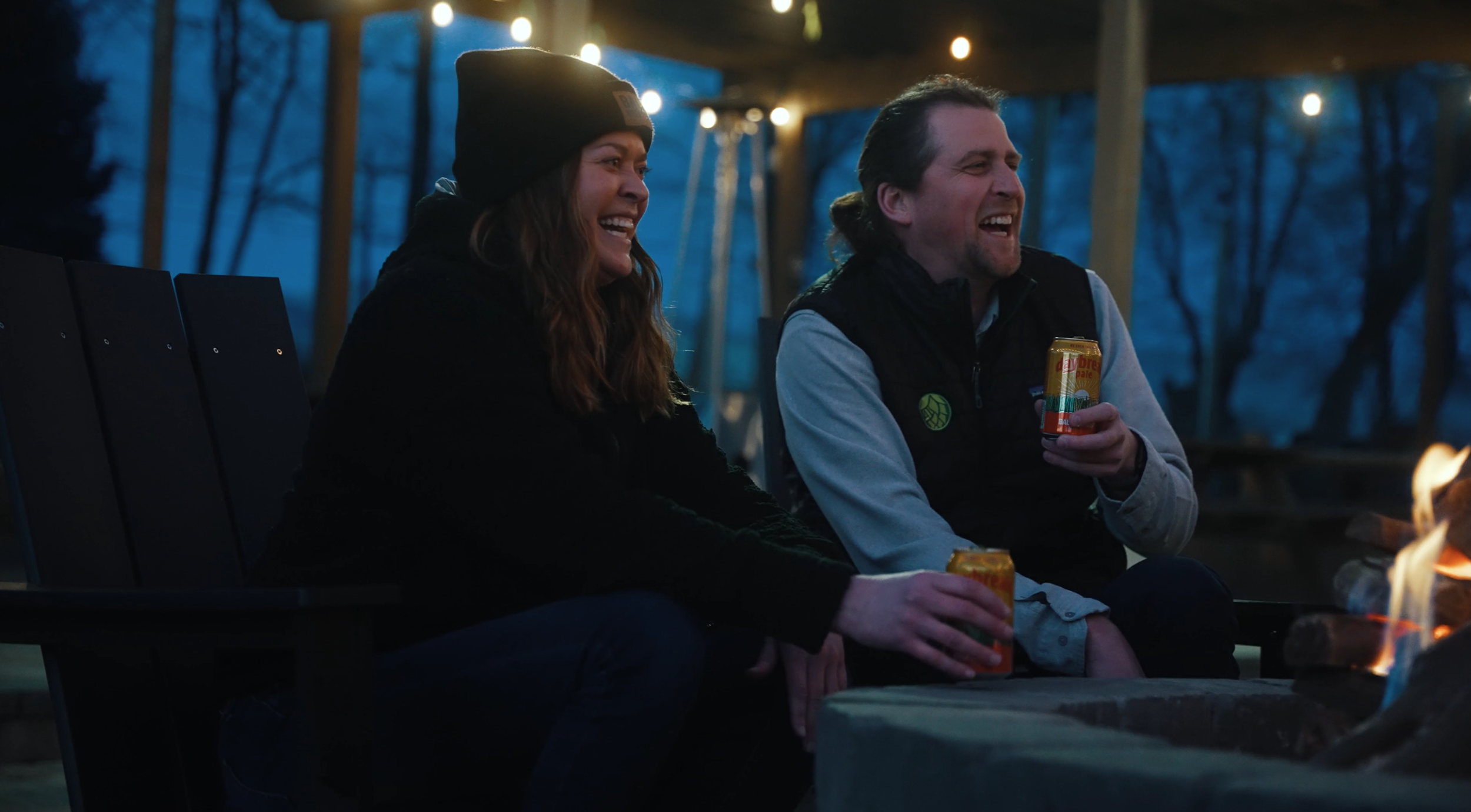Two people sitting on a park bench at dusk, smiling and laughing, holding cans of beverages, with a small fire in front of them and string lights hanging above, surrounded by trees.
