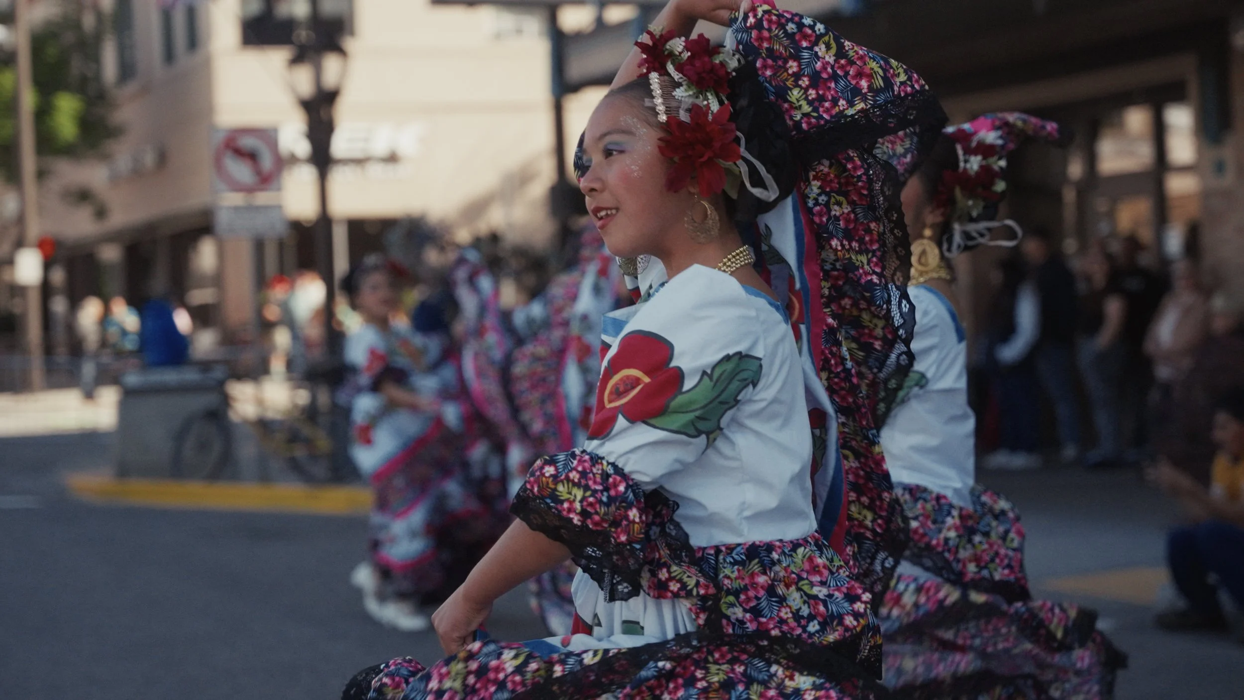 A woman dancing in a street parade, dressed in traditional Mexican attire with floral patterns, a large floral headpiece, and jewelry, smiling as she participates in the celebration.